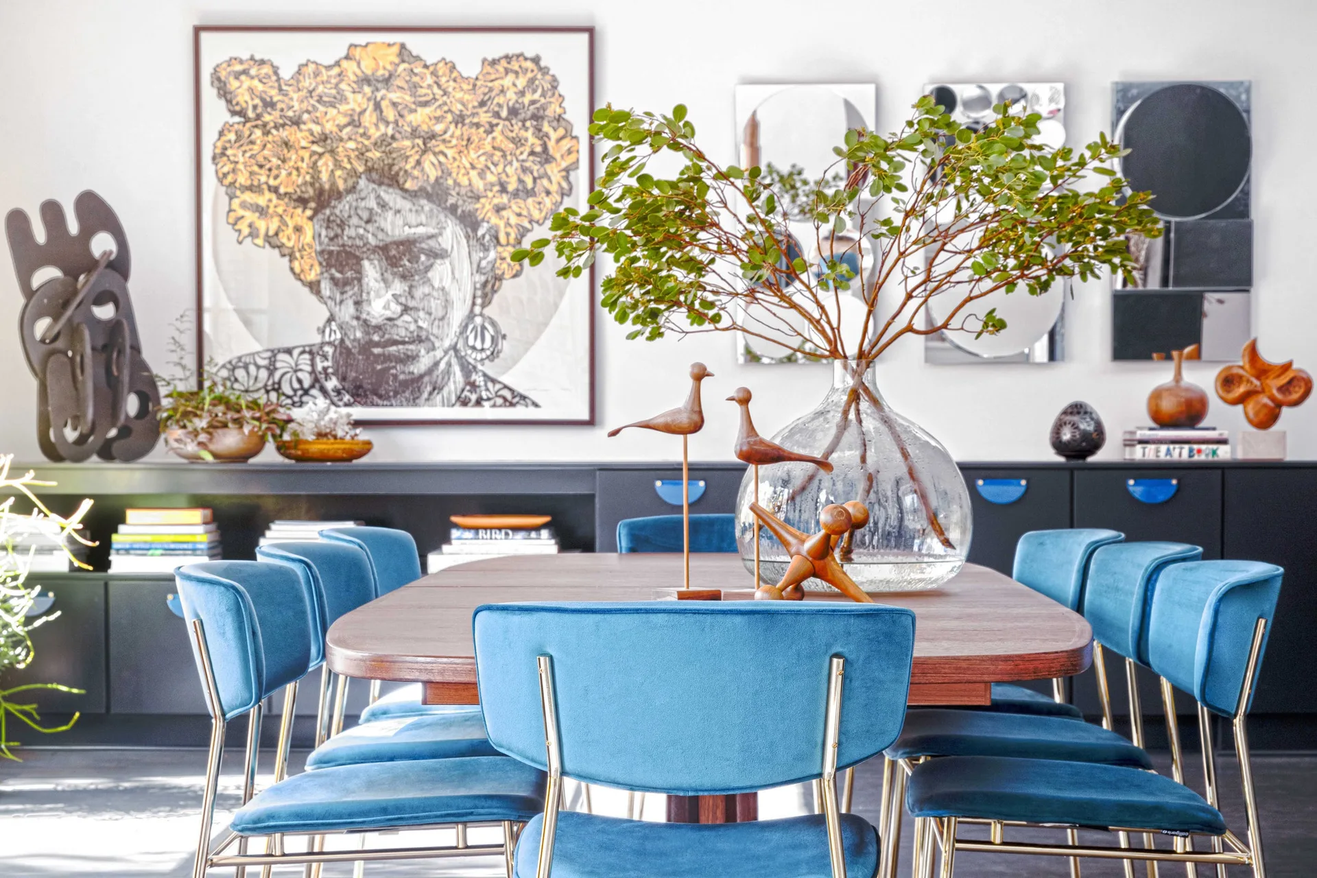 A dining room features a dark wood table with blue velvet chairs, a large glass vase with branches, and a long black sideboard against a white wall.