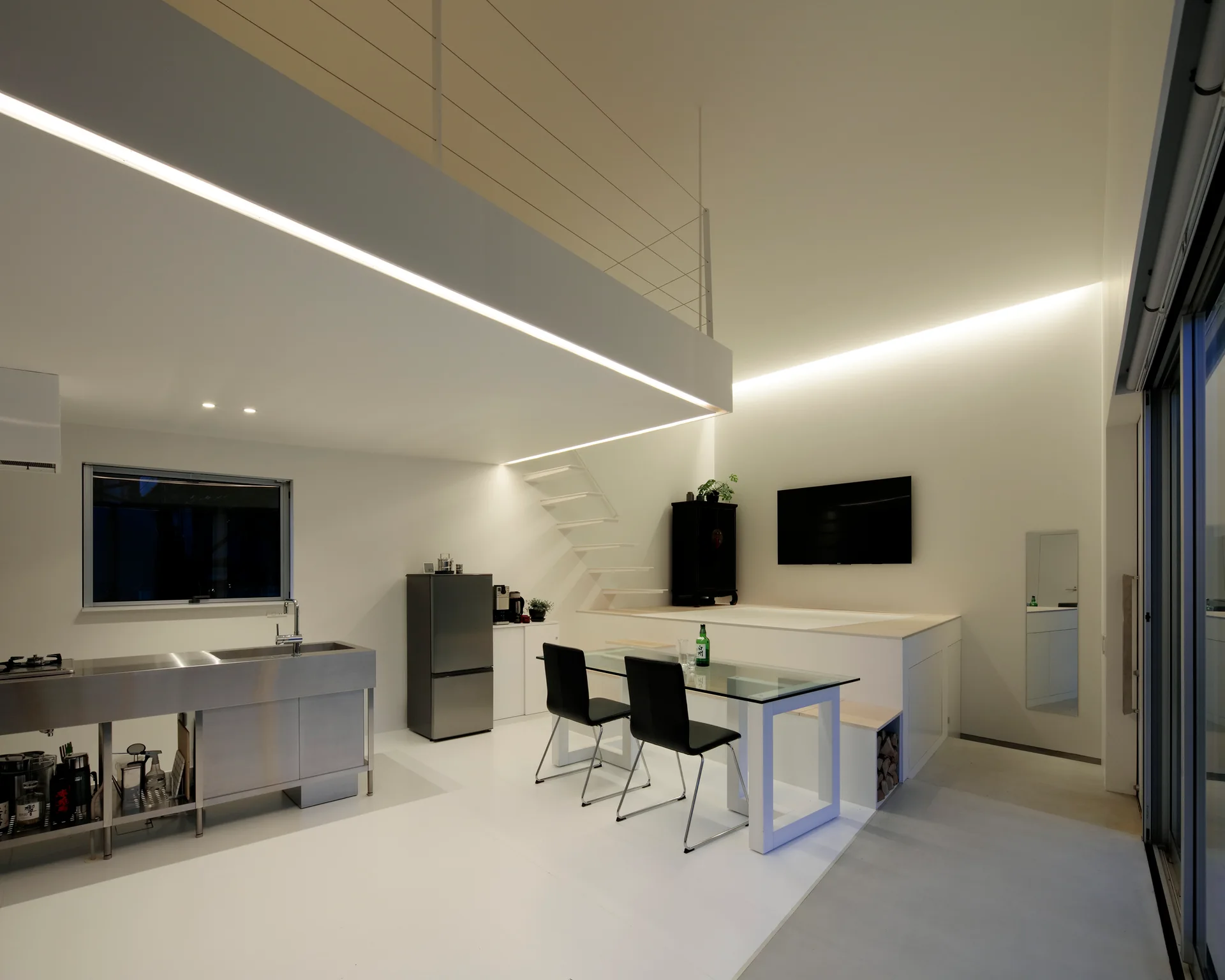 A minimalist white loft featuring a stainless steel kitchen, glass dining table, floating staircase, and mezzanine level with integrated LED lighting.