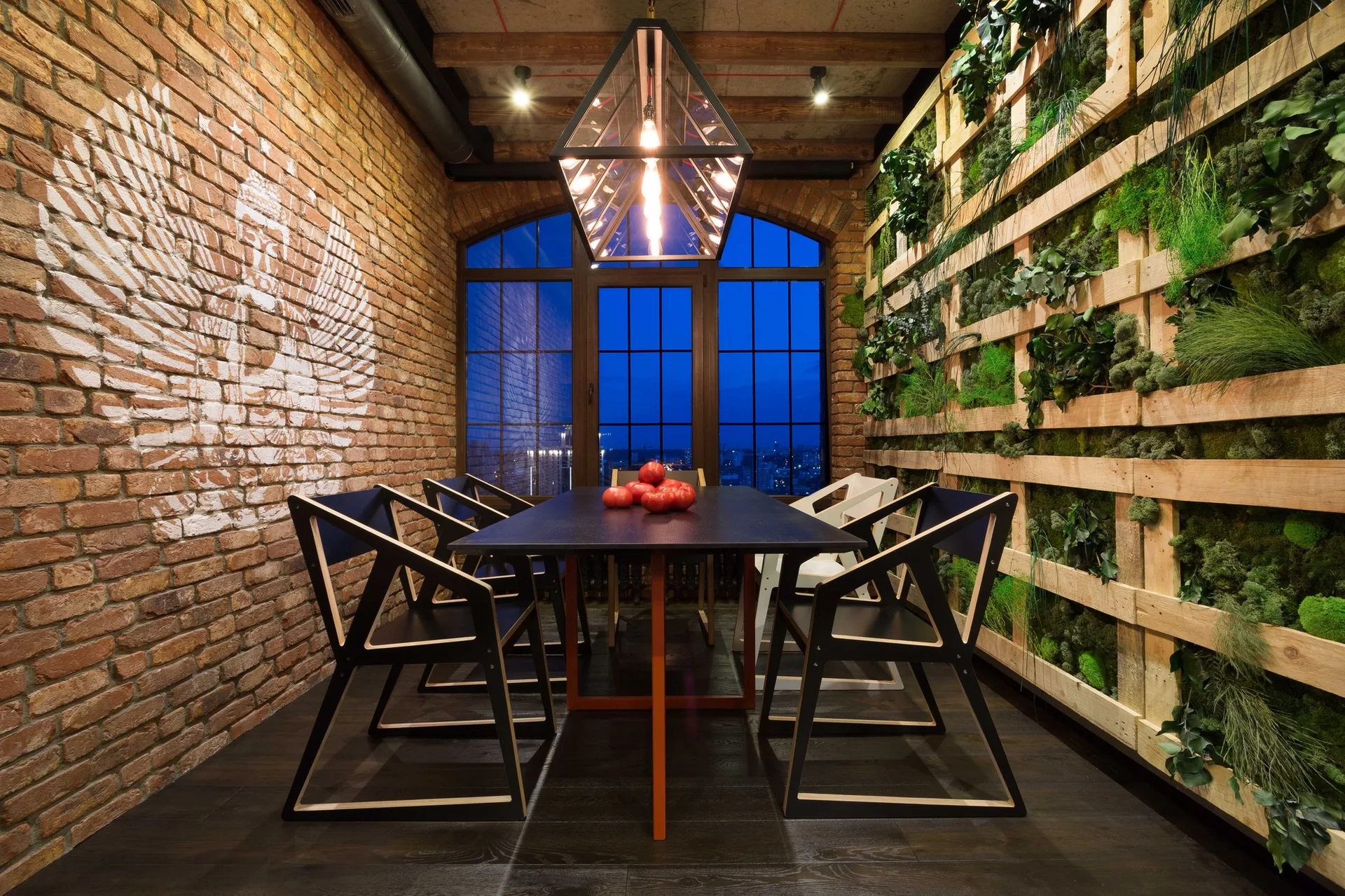 An industrial dining room features a black table with geometric chairs between an exposed brick wall and a wooden vertical moss garden.