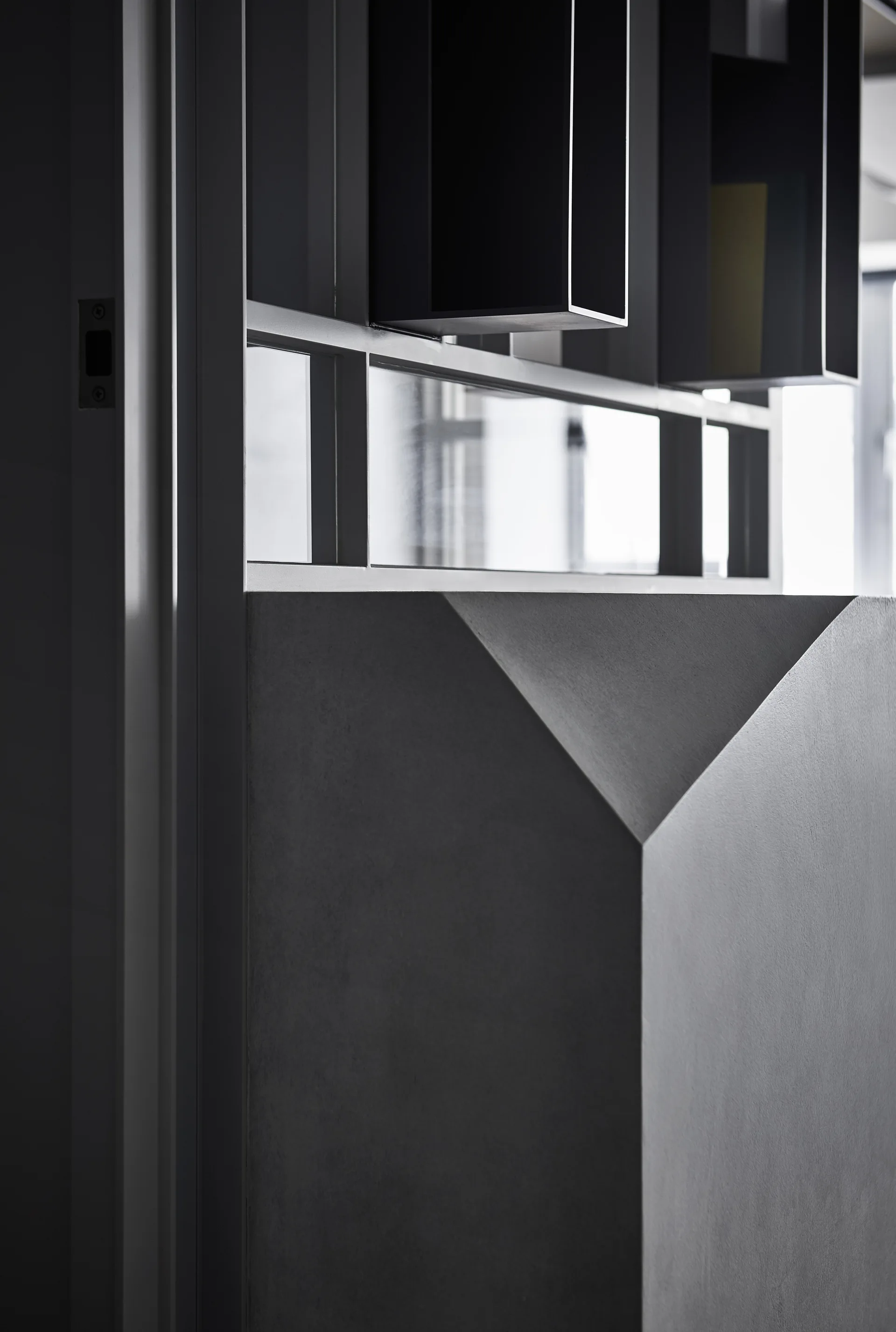 A close-up of a matte gray architectural partition wall with a faceted corner below a glass transom window and black box shelving.