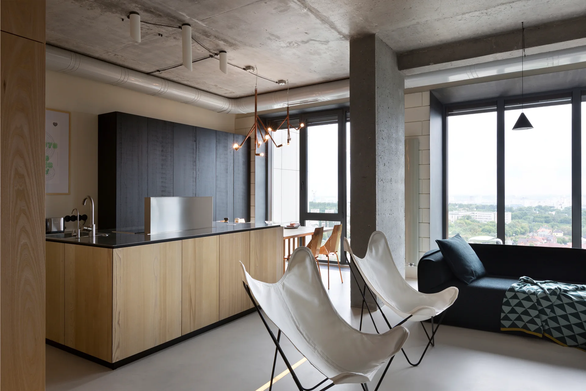An industrial kitchen and living space features a wooden island, raw concrete ceiling, two white butterfly chairs, and large black-framed windows.