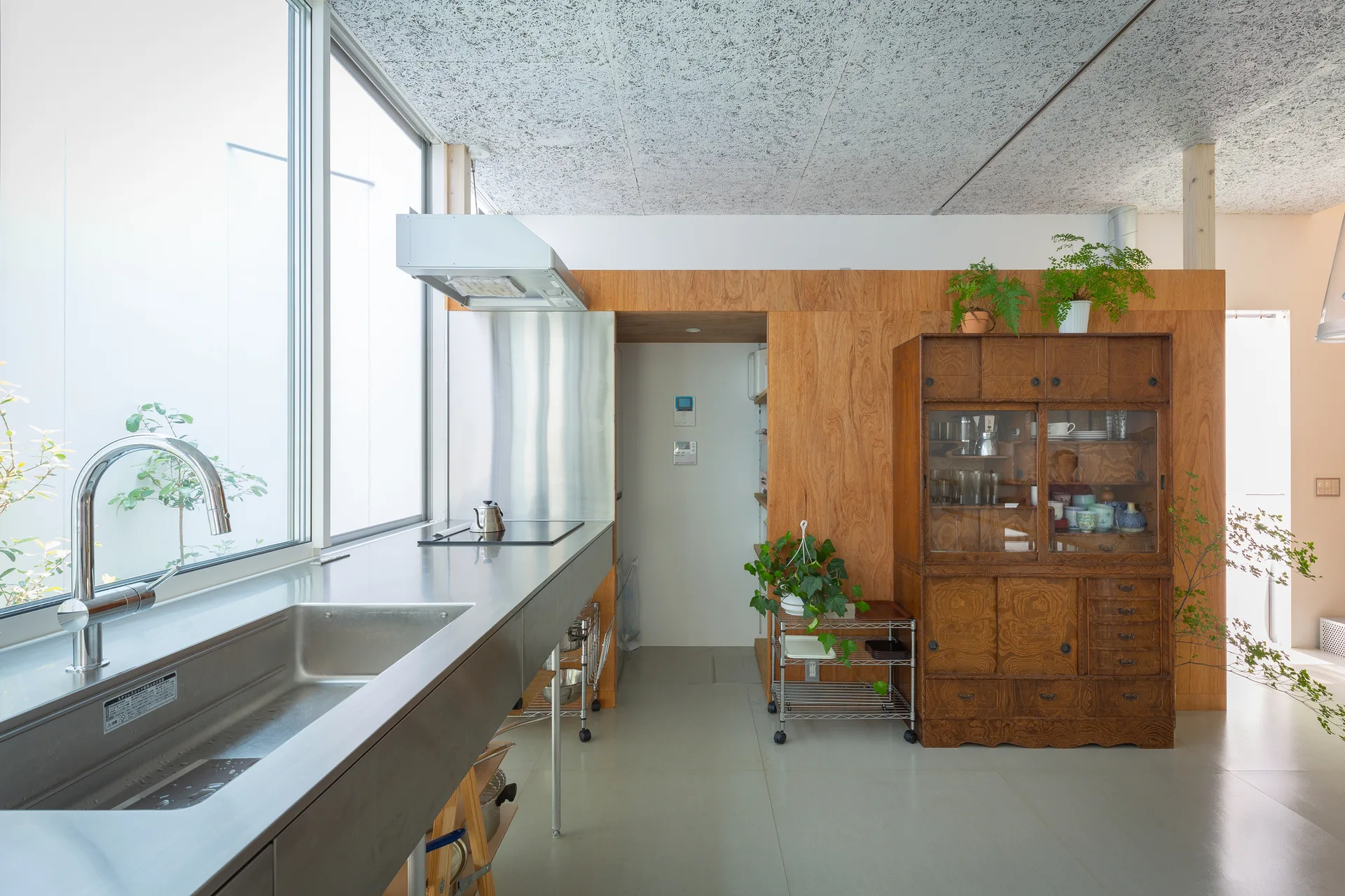 A modern minimalist kitchen with a long stainless steel counter, a large window, a warm wood-paneled wall, and a vintage dark wood cabinet.