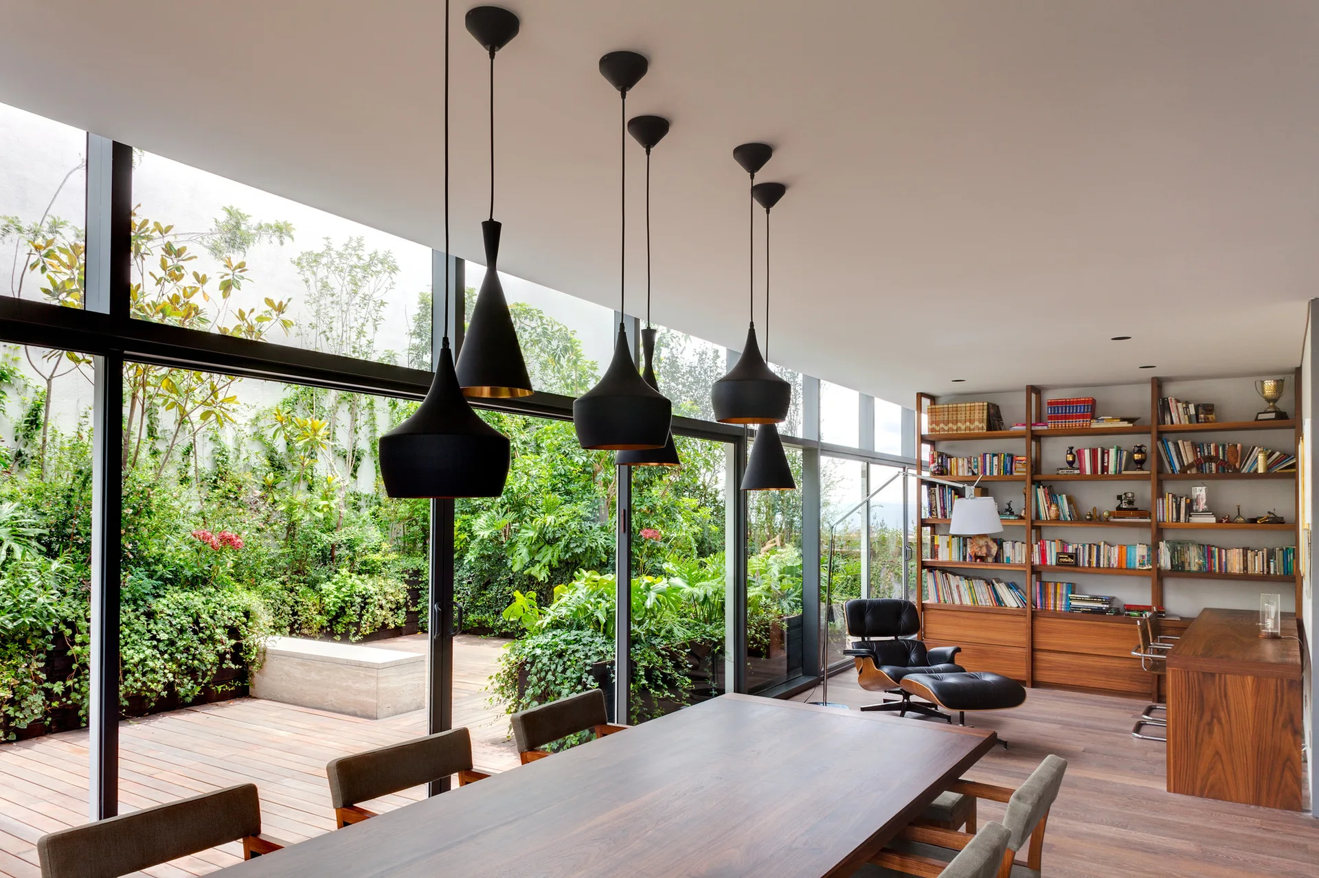 A contemporary dining room with a long wood table, black pendant lights, and a floor-to-ceiling glass wall opening to a garden terrace.