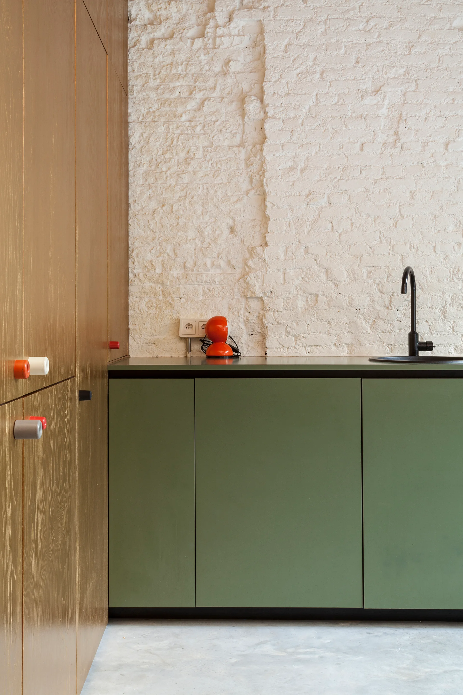 A kitchen detail shows olive green base cabinets, a black sink faucet, and a white painted exposed brick wall next to natural wood tall cabinets.