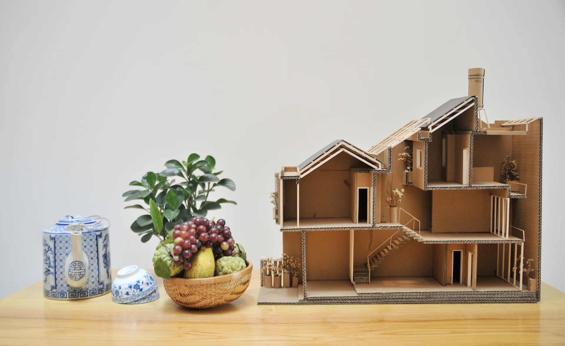 A detailed cardboard architectural house model stands on a light wood table next to a fruit basket and blue-and-white porcelain tableware.