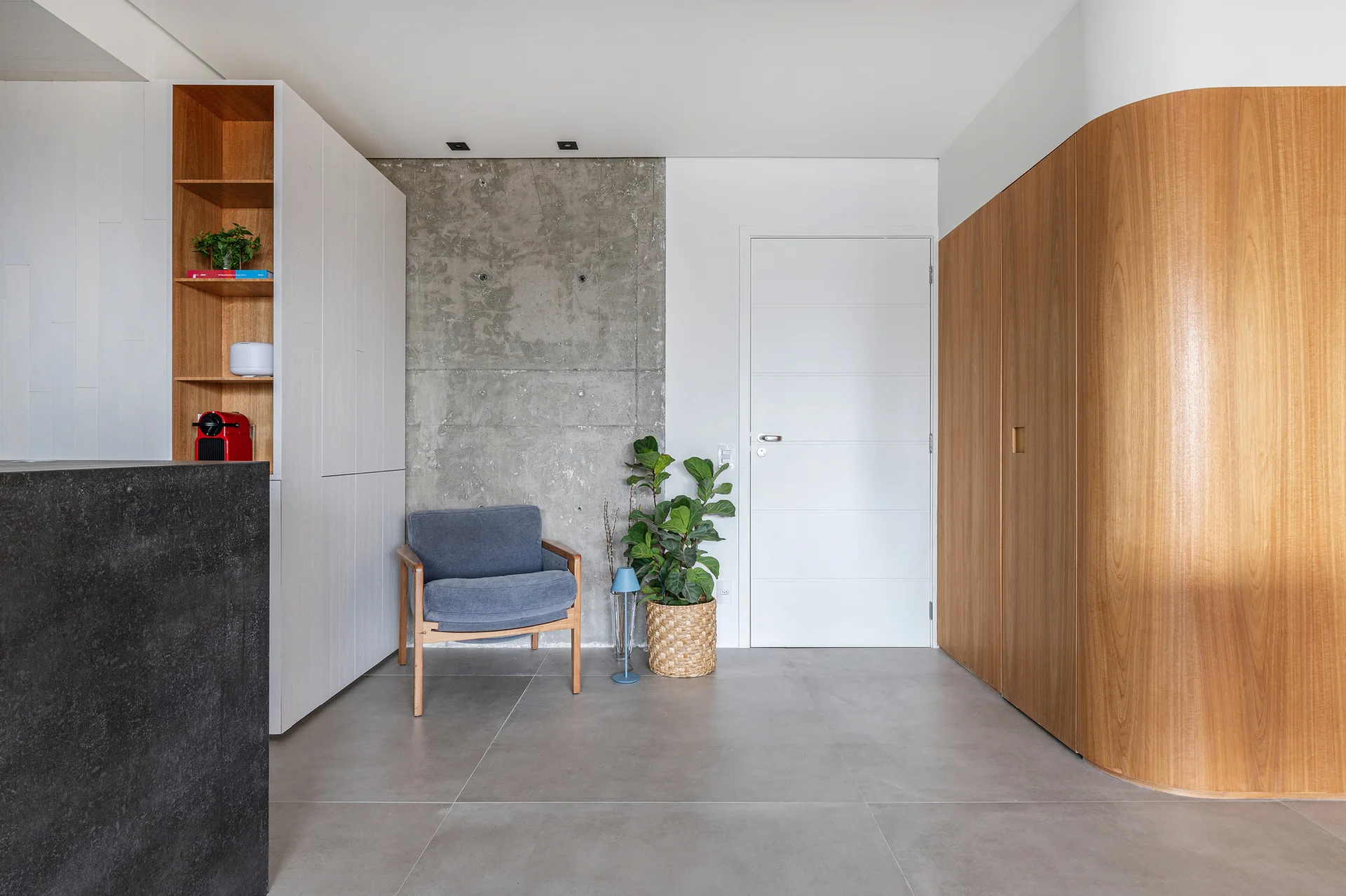 A modern entryway features curved wood wall paneling, an exposed concrete accent wall, a blue armchair, and large grey floor tiles.