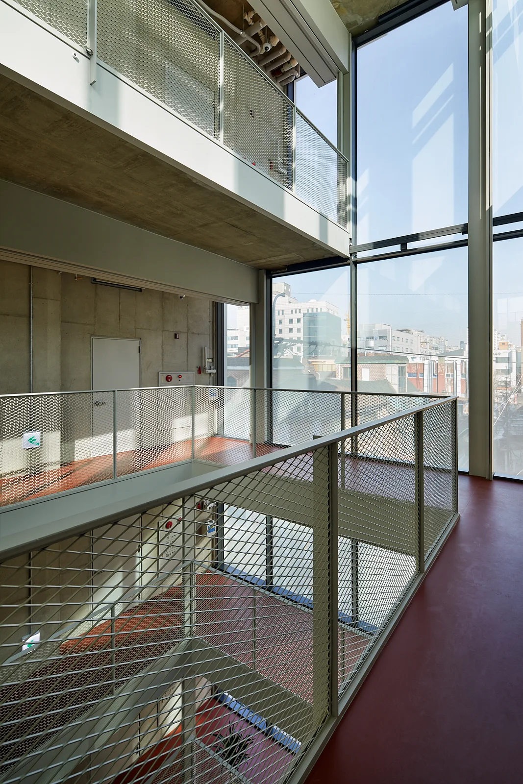 An industrial multi-level interior featuring raw concrete walls, matte red flooring, and light grey metal mesh railings alongside large glass windows.