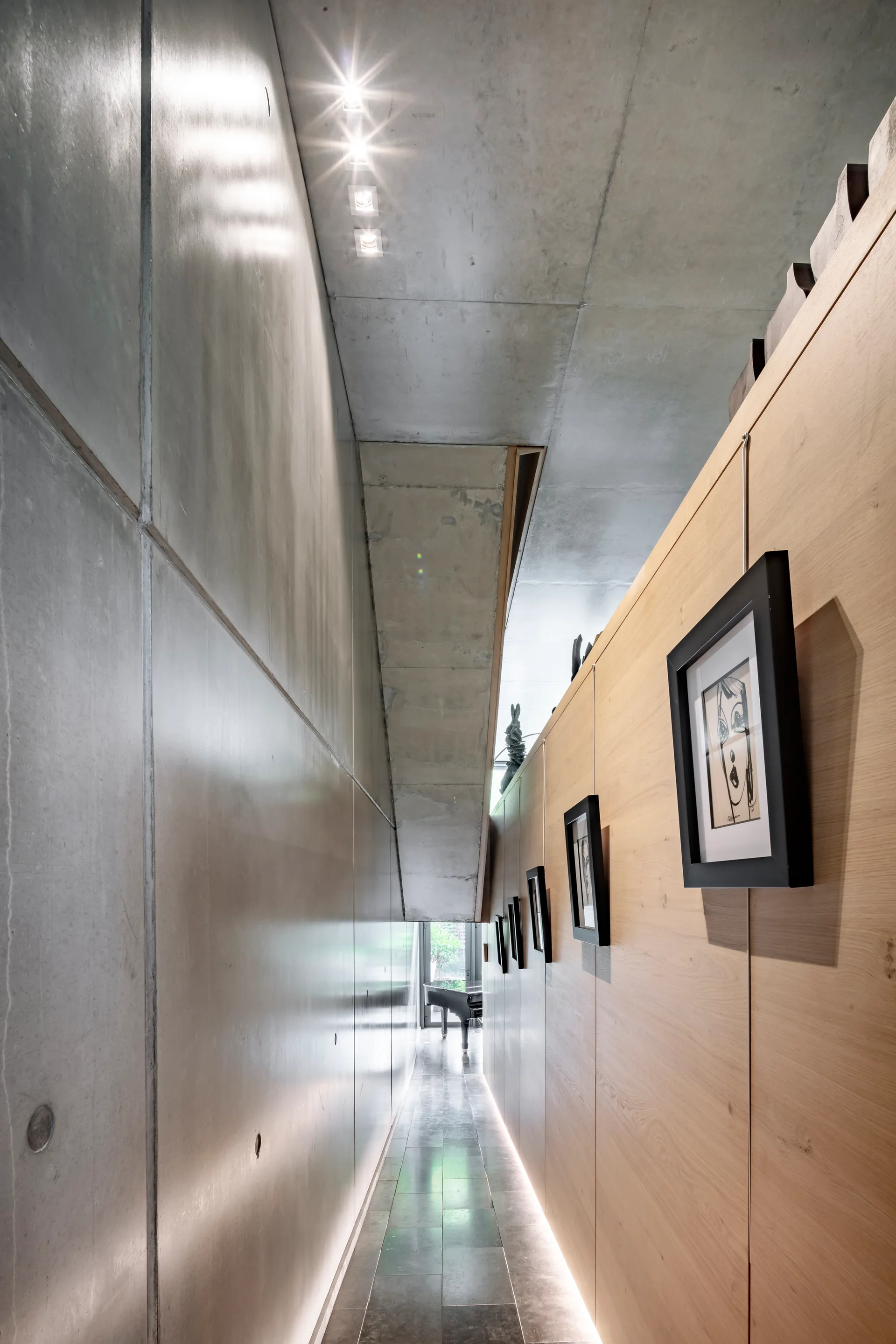 A narrow hallway features a smooth concrete wall on one side, light wood paneling with framed art on the other, and a concrete ceiling with spotlights.