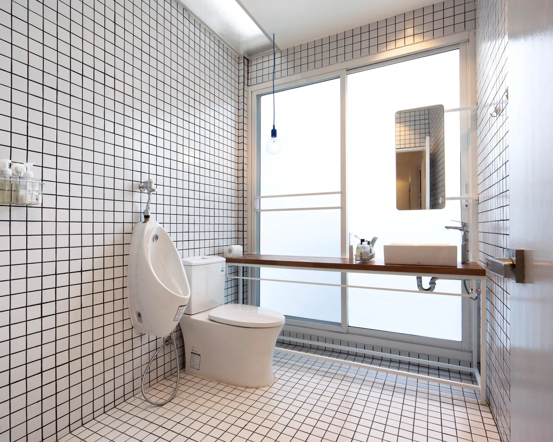 A minimalist white bathroom features walls and floors covered in small square grid tiles, a wall-mounted urinal, and a slim wood shelf vanity.