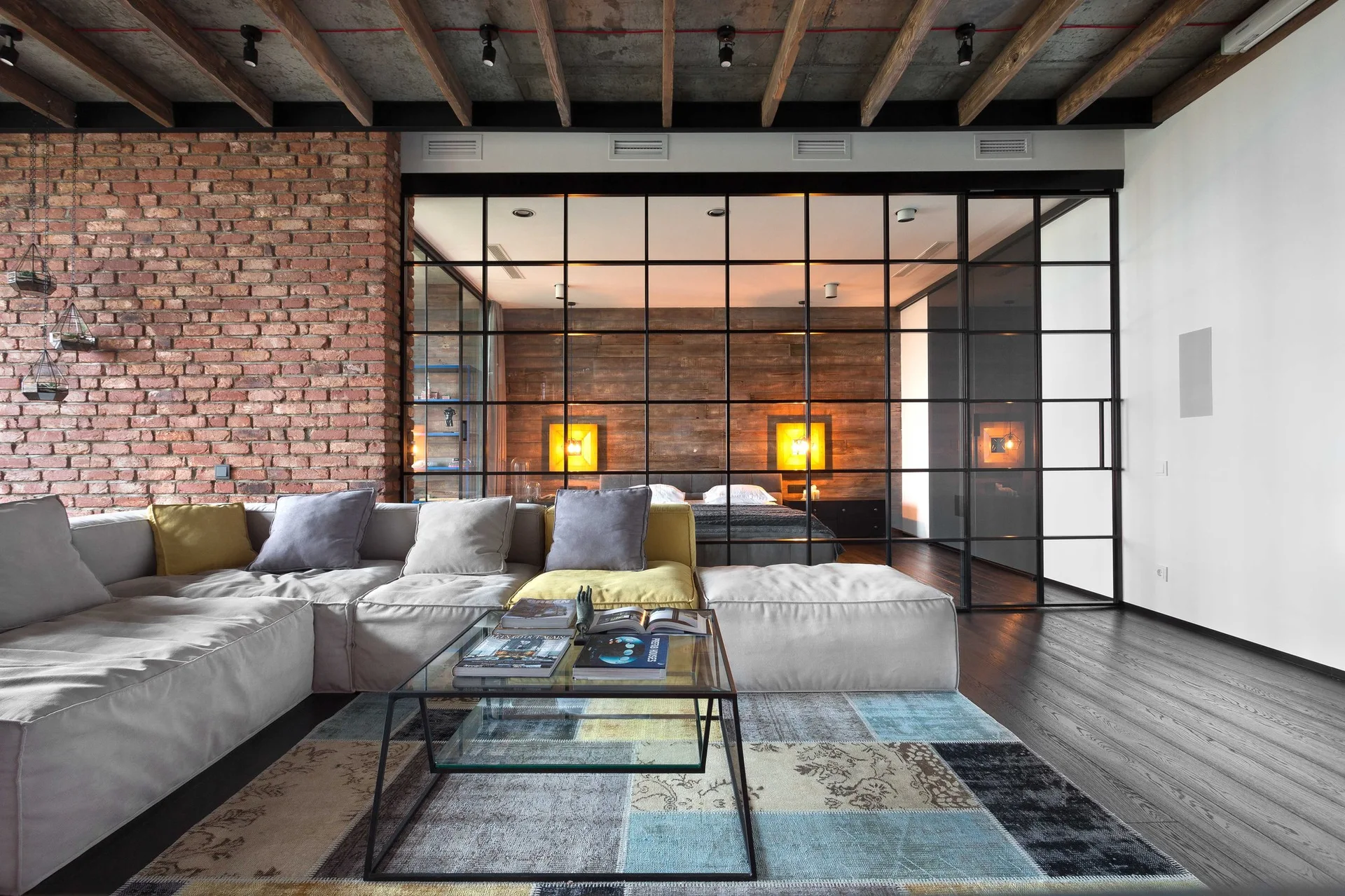 An industrial living room features a modular sectional sofa, a patchwork rug, an exposed brick wall, and a black metal glass grid partition.