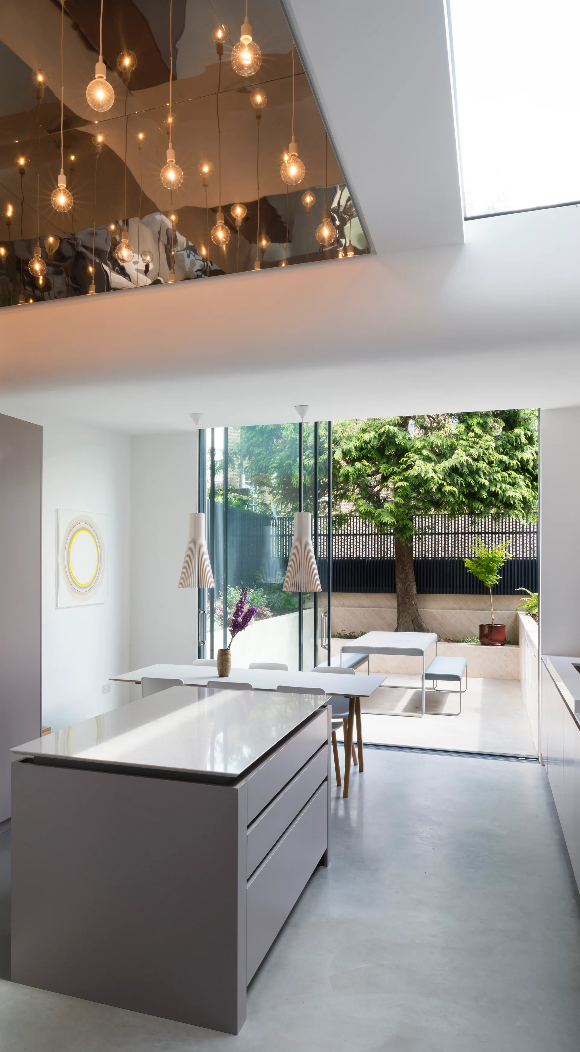 A modern minimalist kitchen features a grey island, a white dining table under a mirrored ceiling, and large glass doors opening to an outdoor patio.