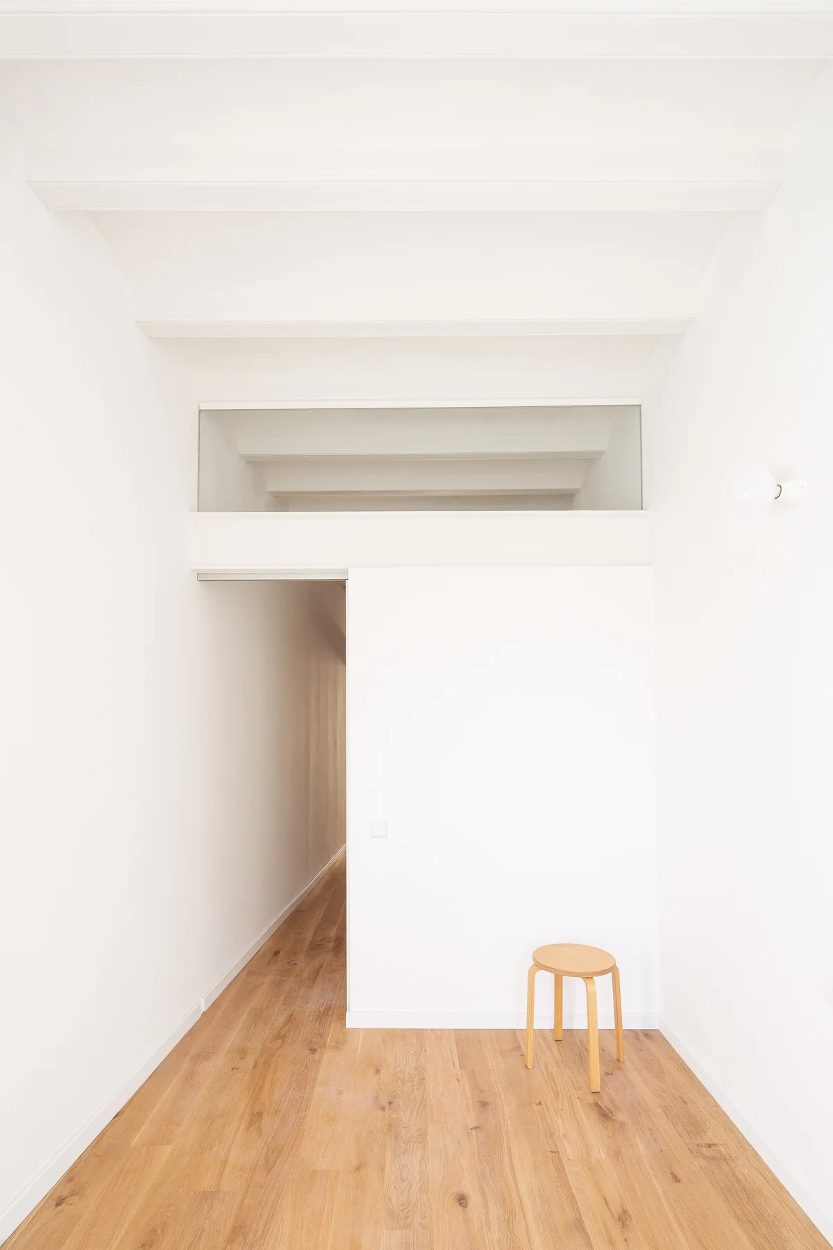 A minimalist white hallway features light oak flooring, a wide partition doorway with a glass transom window, and a single three-legged wooden stool.
