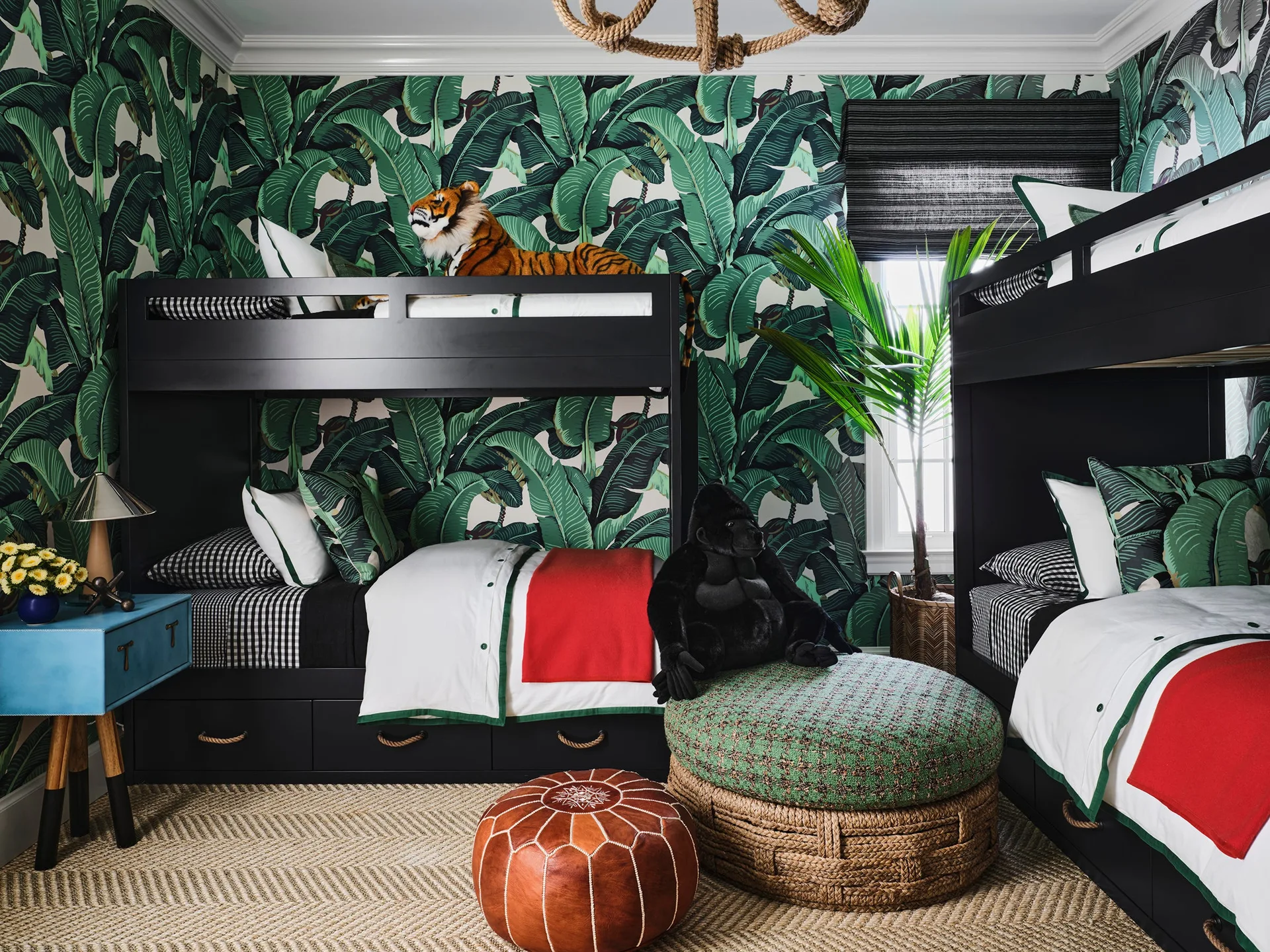 A children's bedroom features black wood bunk beds against green palm leaf wallpaper, a herringbone fiber rug, and a leather pouf.