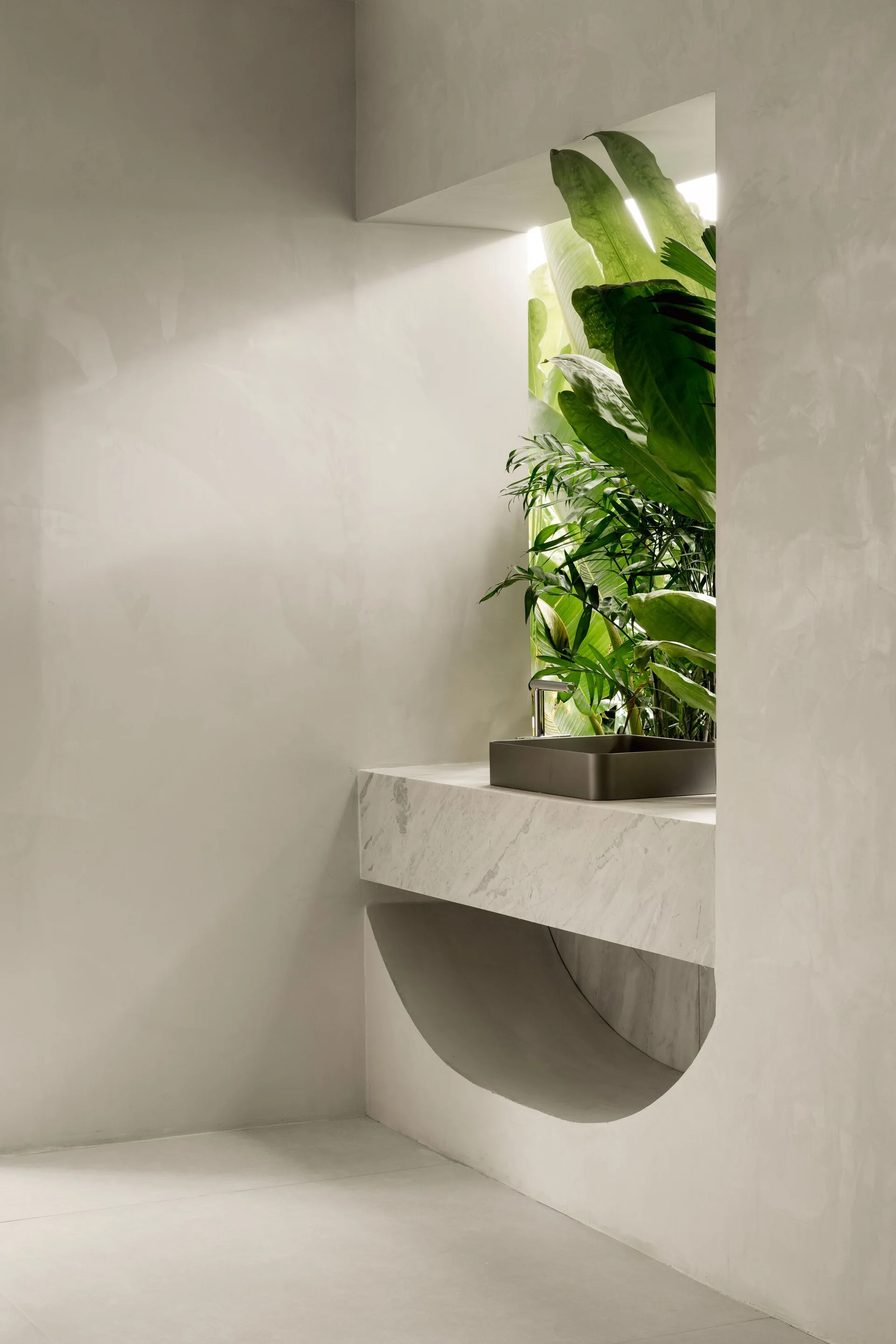 A minimalist bathroom features a floating marble vanity with a dark sink set in a recessed plaster wall niche filled with lush green plants.