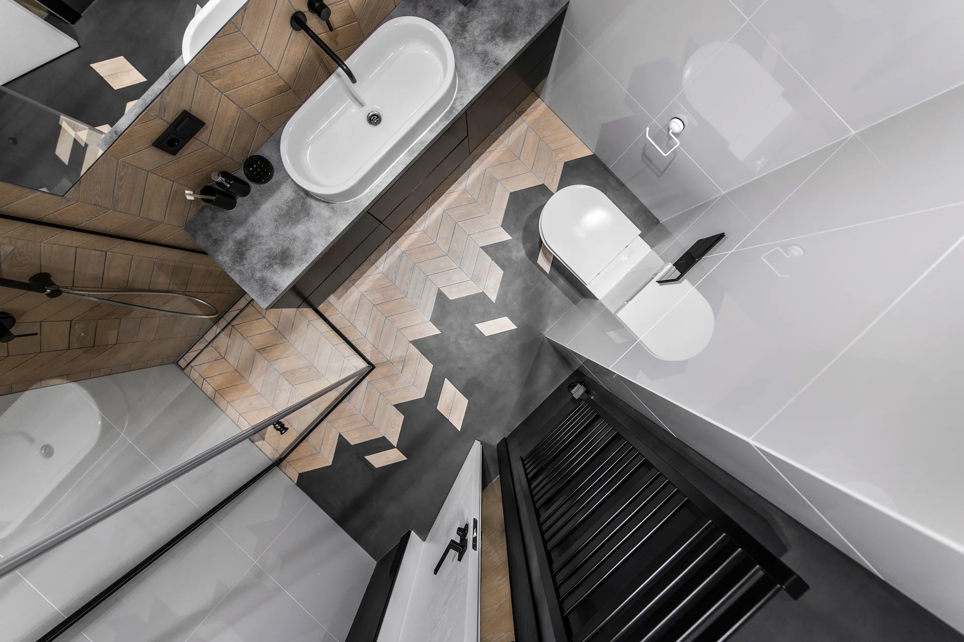 An overhead view of a bathroom showing a chevron tile floor transitioning into dark gray tile beside a dark gray vanity and white wall-hung toilet.