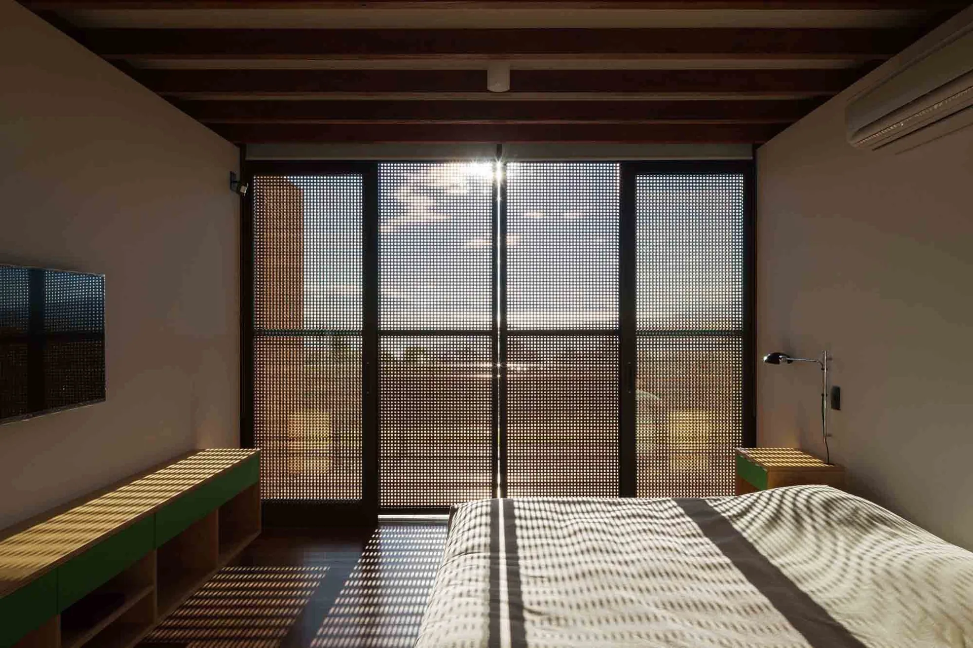 A modern bedroom features exposed ceiling beams and sliding glass doors covered with a grid-patterned screen filtering sunlight onto the floor.