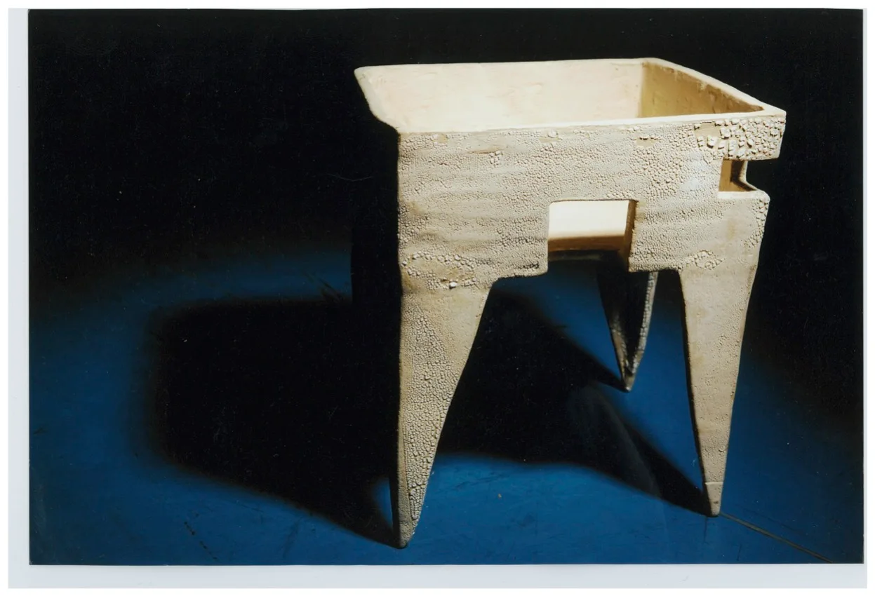 An off-white sculptural ceramic side table with tapered legs and cutout details sits on a dark blue surface against a black background.