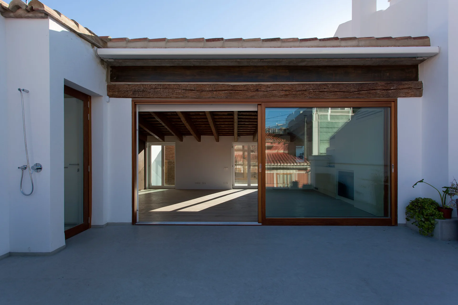 A concrete terrace features large wooden-frame sliding glass doors beneath a rustic wood beam, with a chrome shower fixture mounted to a white exterior wall.