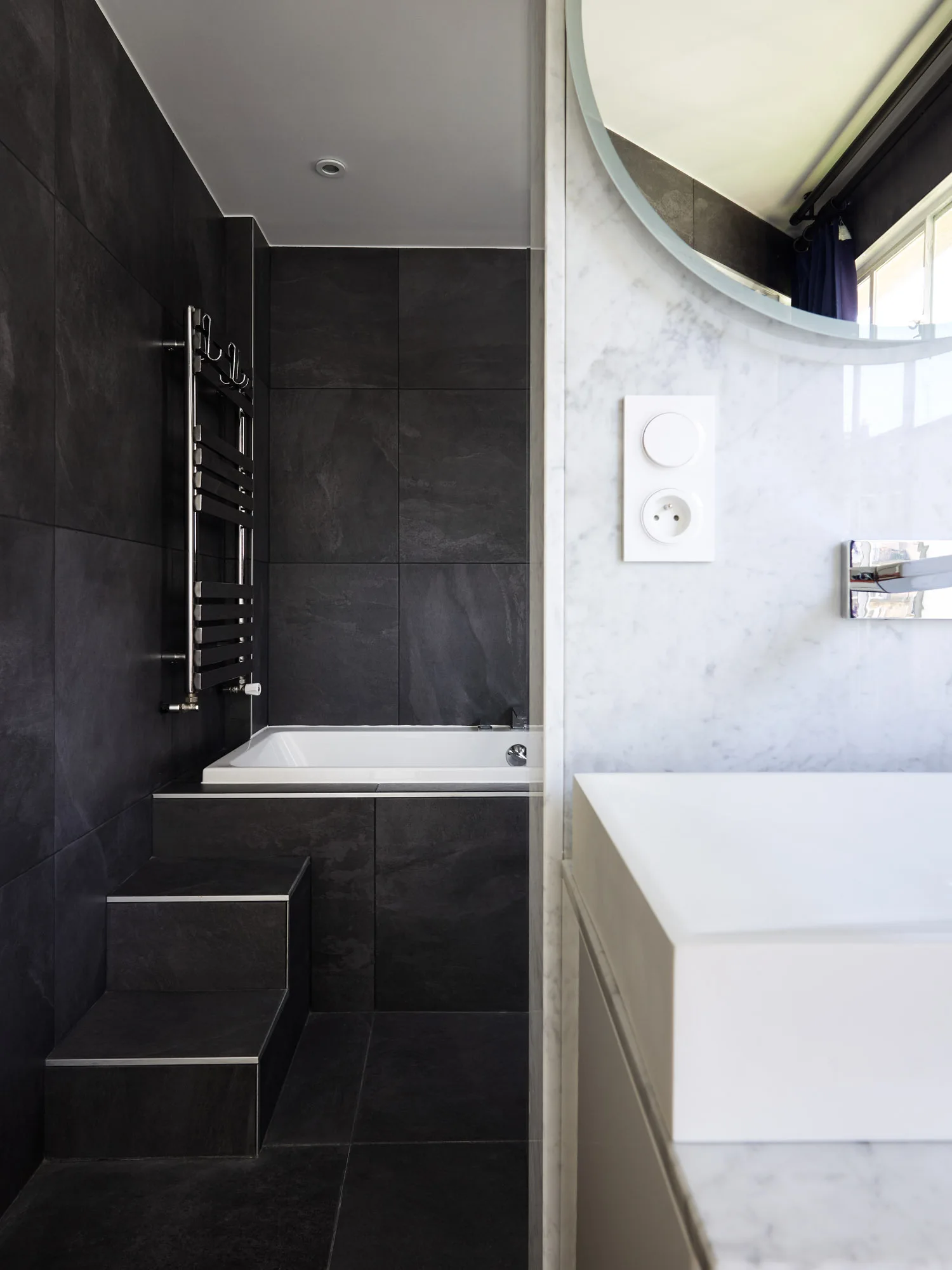 A contemporary bathroom showing a step-up bathtub surrounded by dark stone tiles next to a white marble vanity with a round mirror.