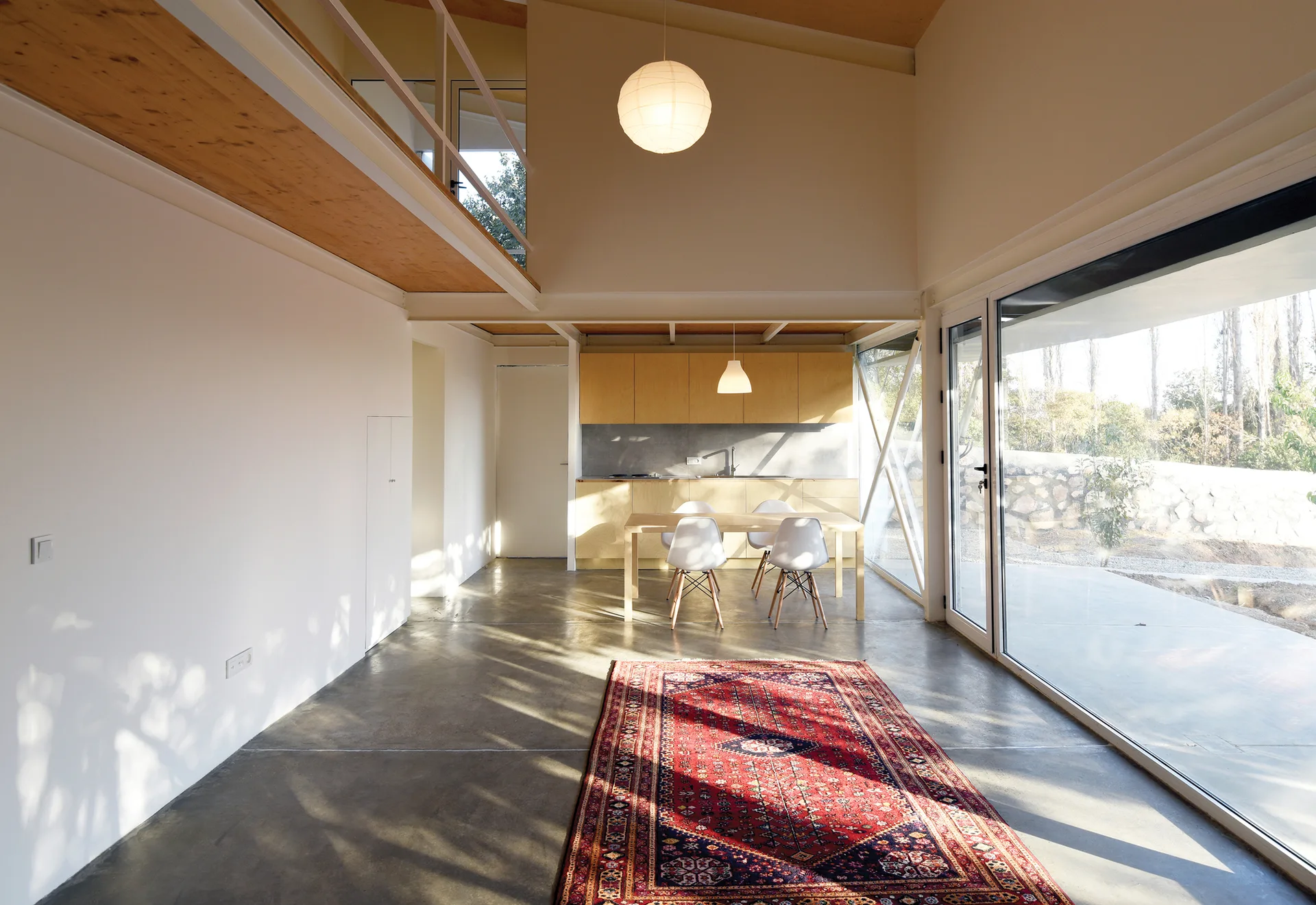 A double-height modern loft featuring a polished concrete floor, a traditional red rug, a dining set, and a light wood kitchen under a mezzanine.