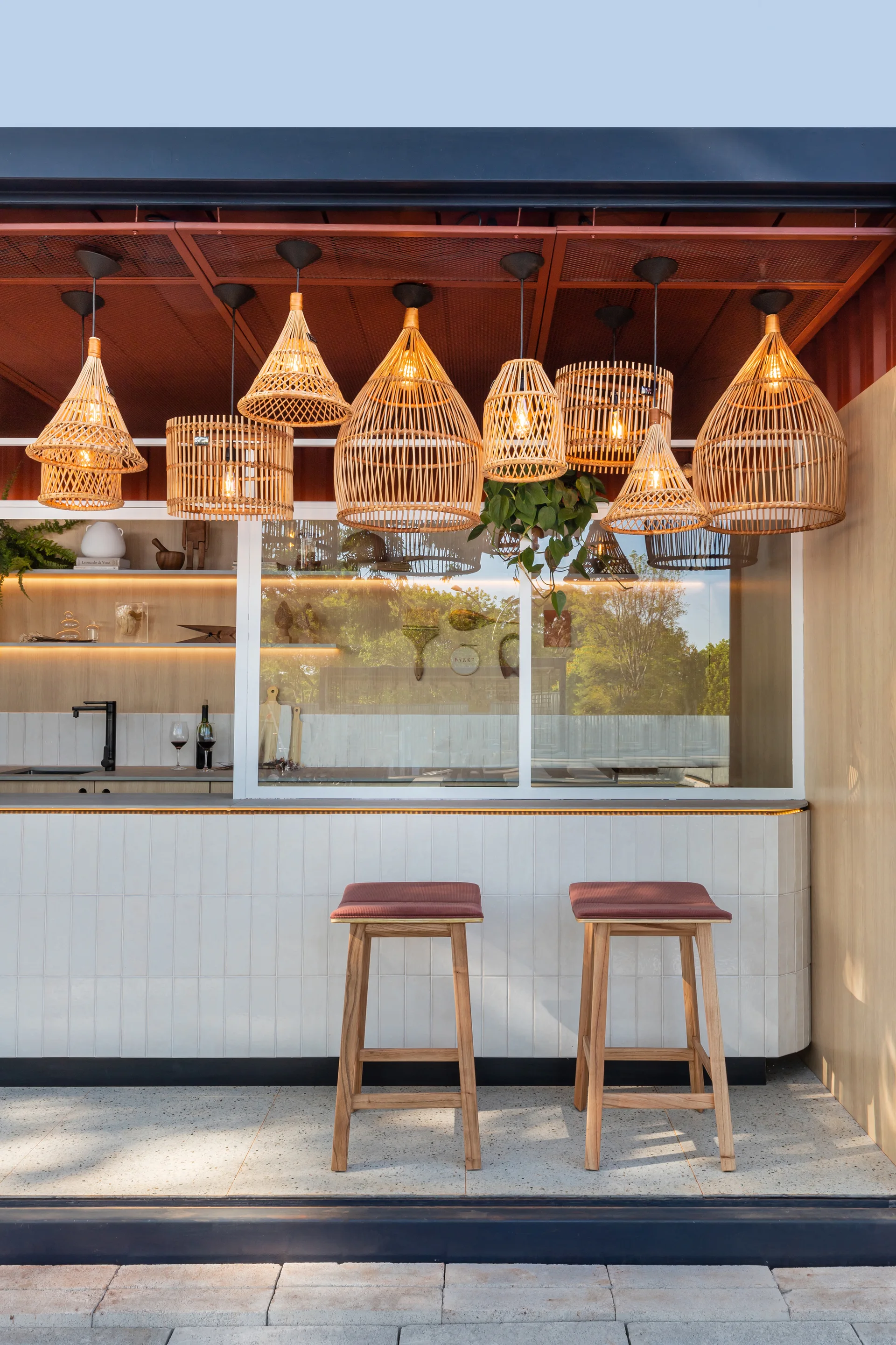 An outdoor bar features a vertically tiled white counter, two wooden bar stools, and a dense cluster of various woven rattan pendant lights hanging from above.