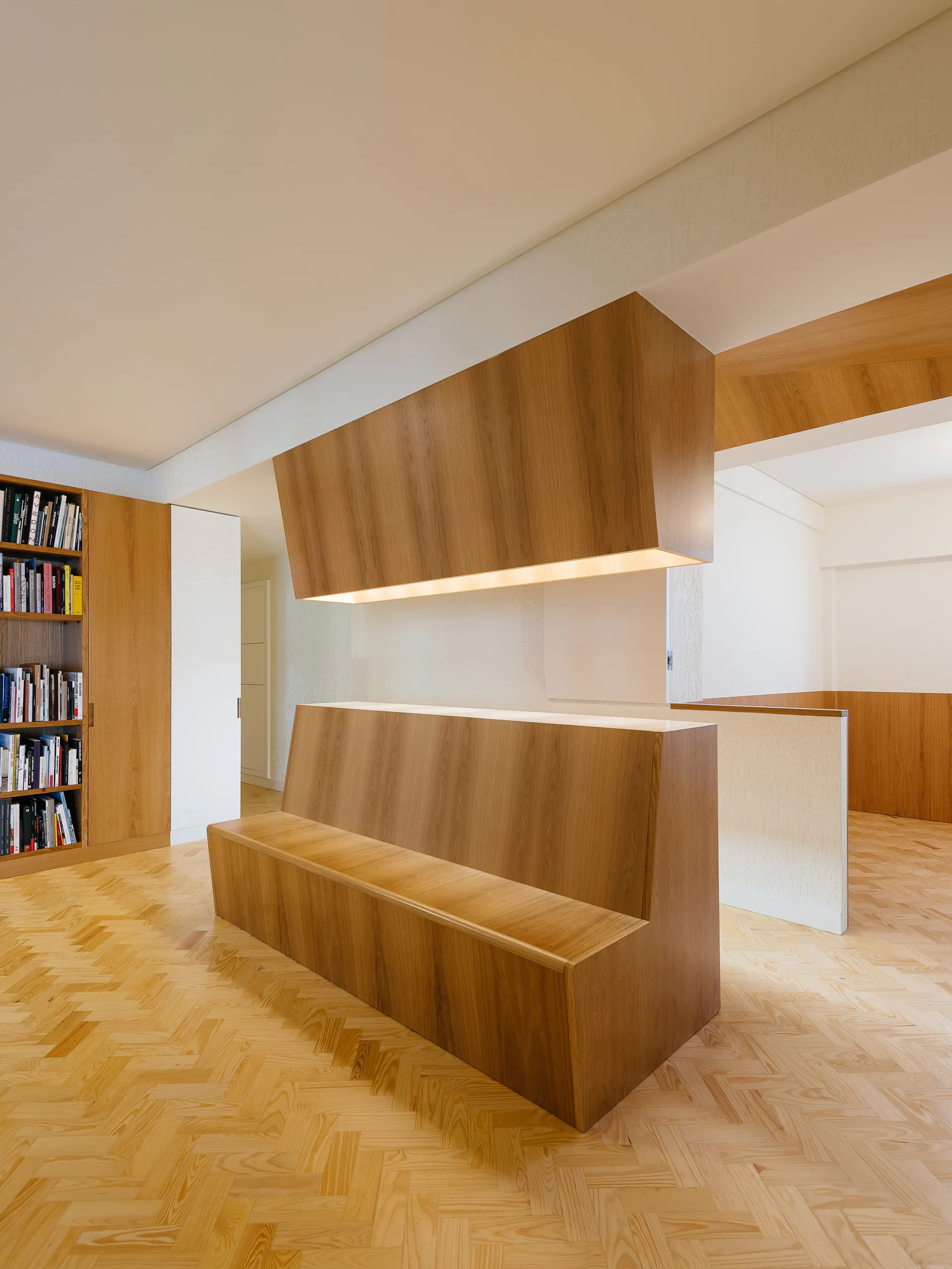 A minimalist hallway features a central custom wooden bench, a matching floating wooden ceiling structure with warm lighting, and herringbone wood floors.