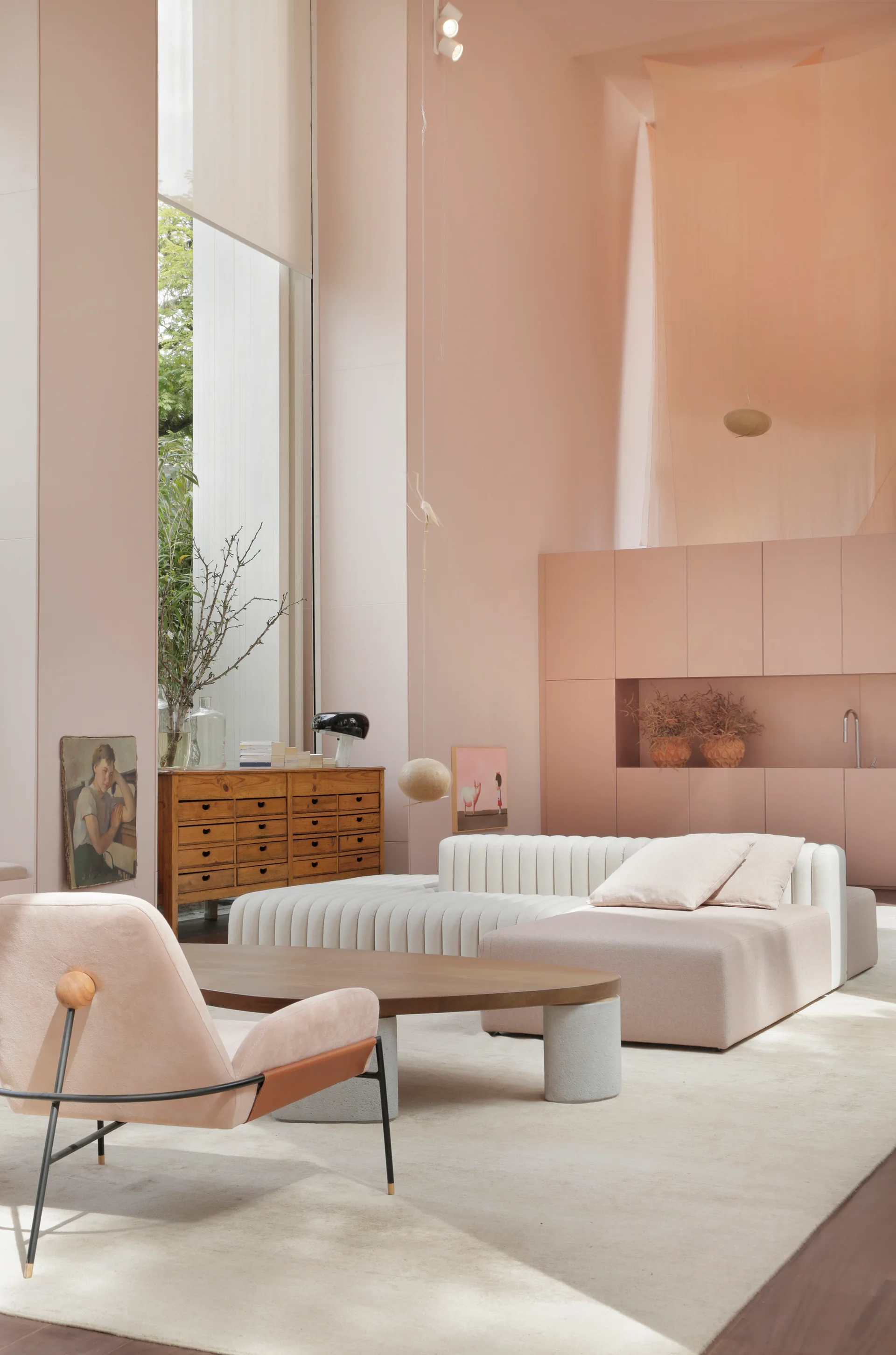 A minimalist living room with pale pink walls, featuring a modular white sofa, a wooden apothecary chest, and a low oval coffee table.