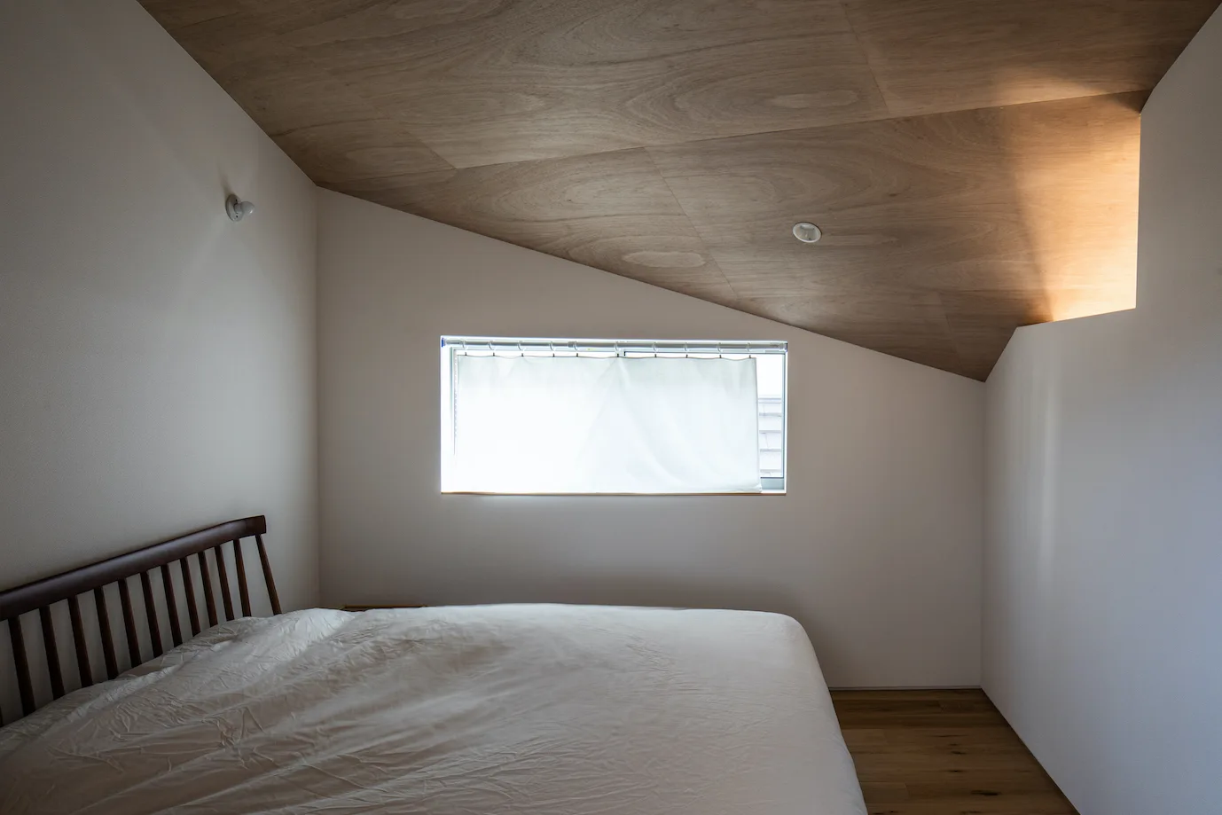 A minimalist bedroom features a bed with a wooden spindle headboard beneath a sloped plywood ceiling and a small window on a plain white wall.