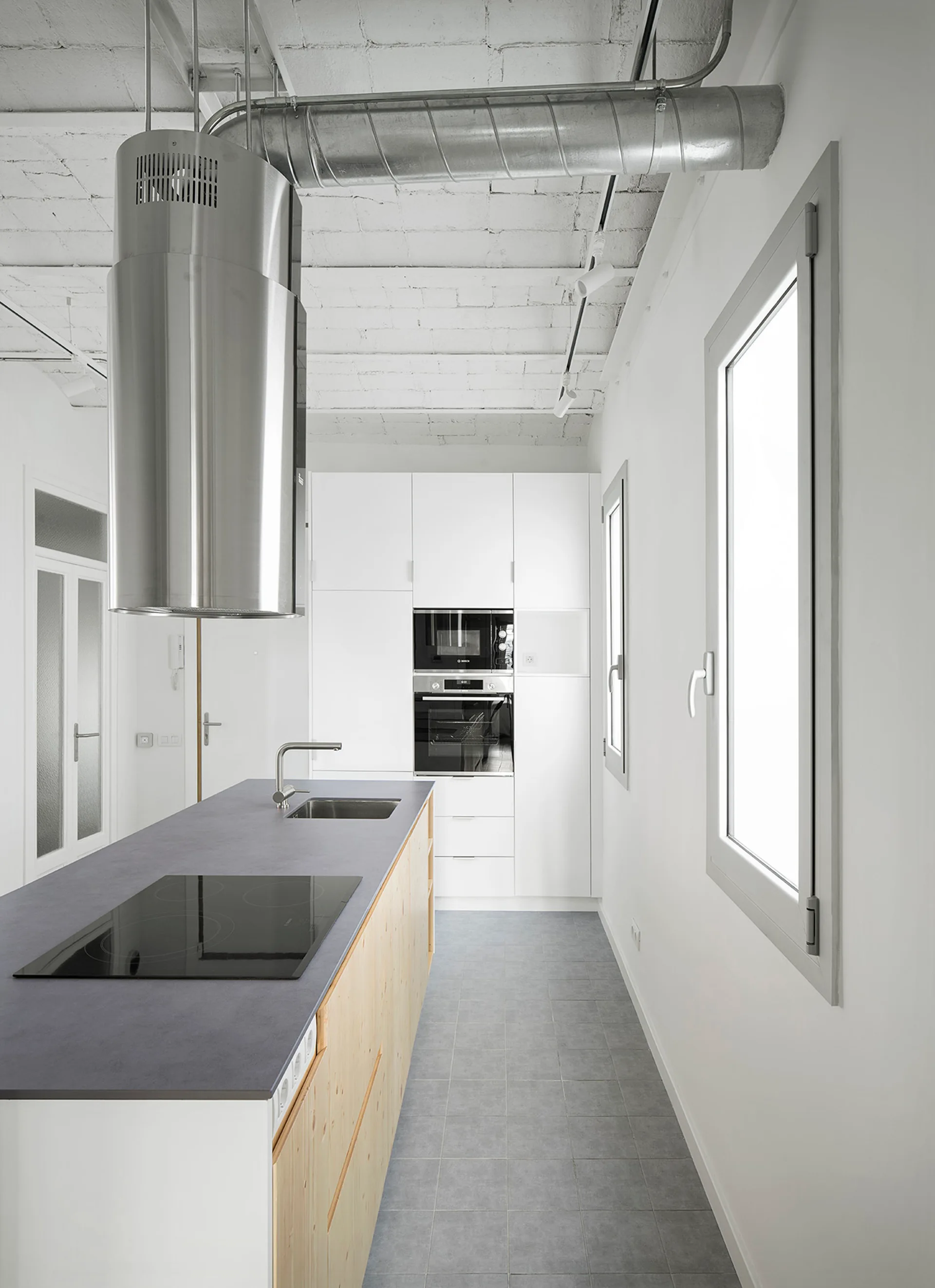 A modern kitchen featuring a central wood island with a dark grey countertop, white cabinetry, and an exposed white brick ceiling.