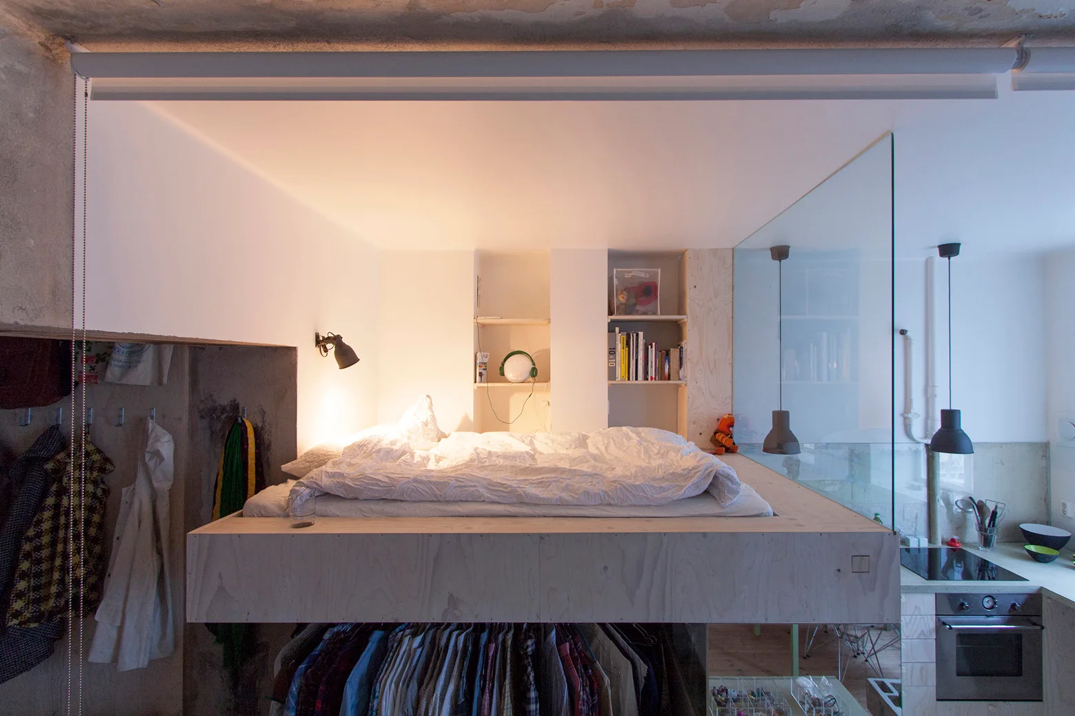 An elevated plywood loft bed platform built above an open closet area with hanging clothes next to a kitchen glass partition.