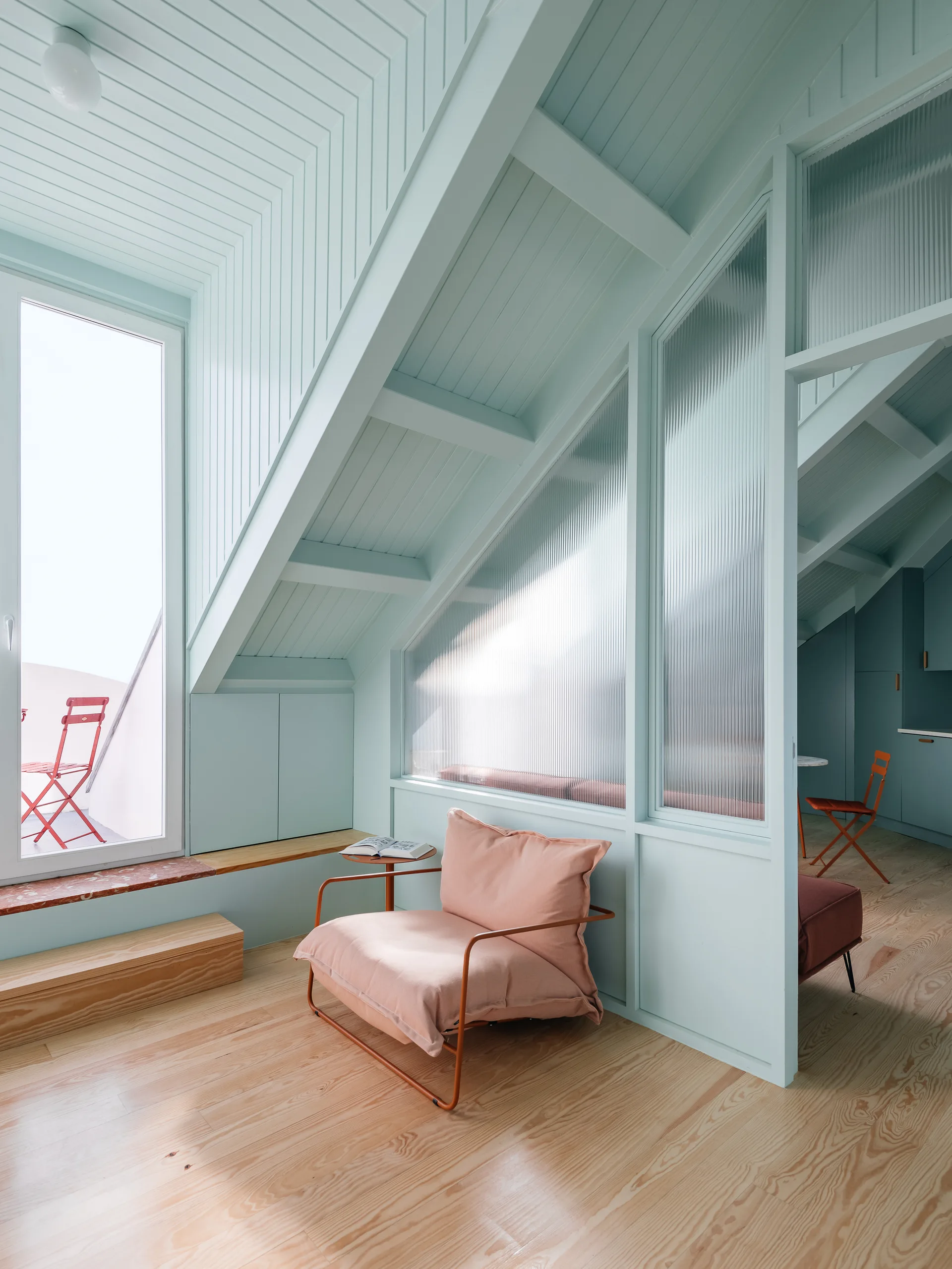 A loft sitting space with mint green paneled sloped ceilings, a soft pink lounge chair, and a fluted glass partition over light wood floors.