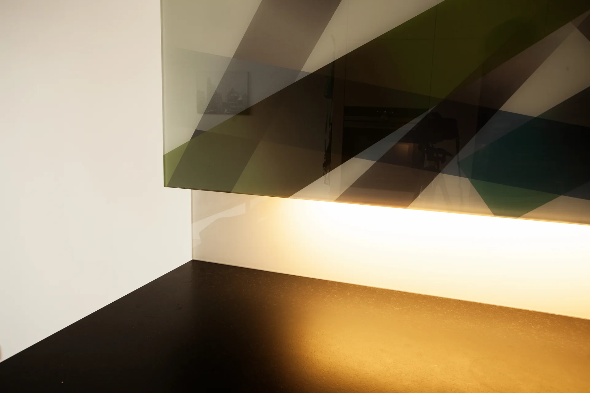 A dark kitchen countertop sits beneath a glossy geometric pattern glass cabinet illuminated by warm integrated under-cabinet lighting.