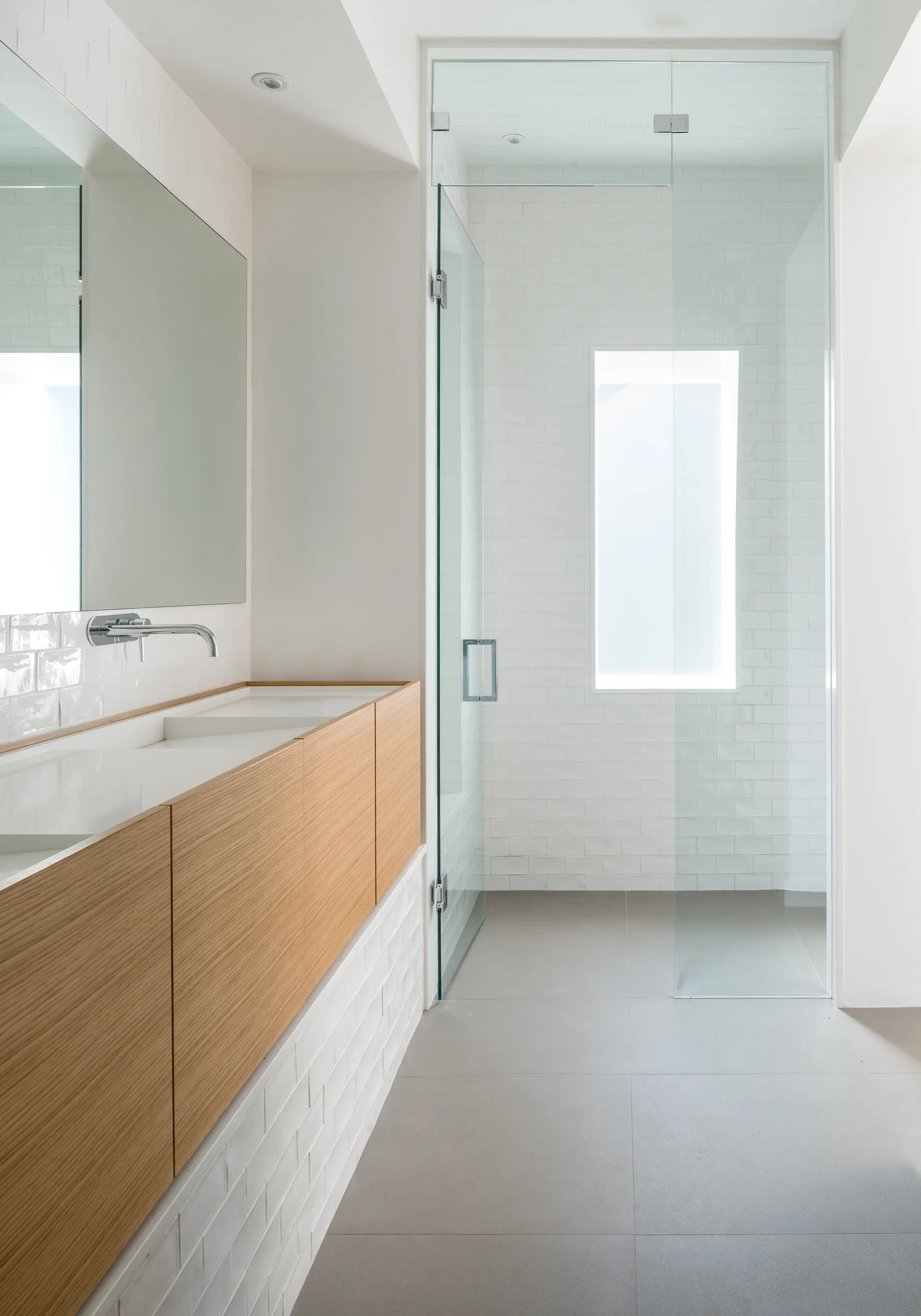 A modern bathroom features a light wood floating vanity with a white countertop, a frameless mirror, and a glass-enclosed shower with white subway tiles.