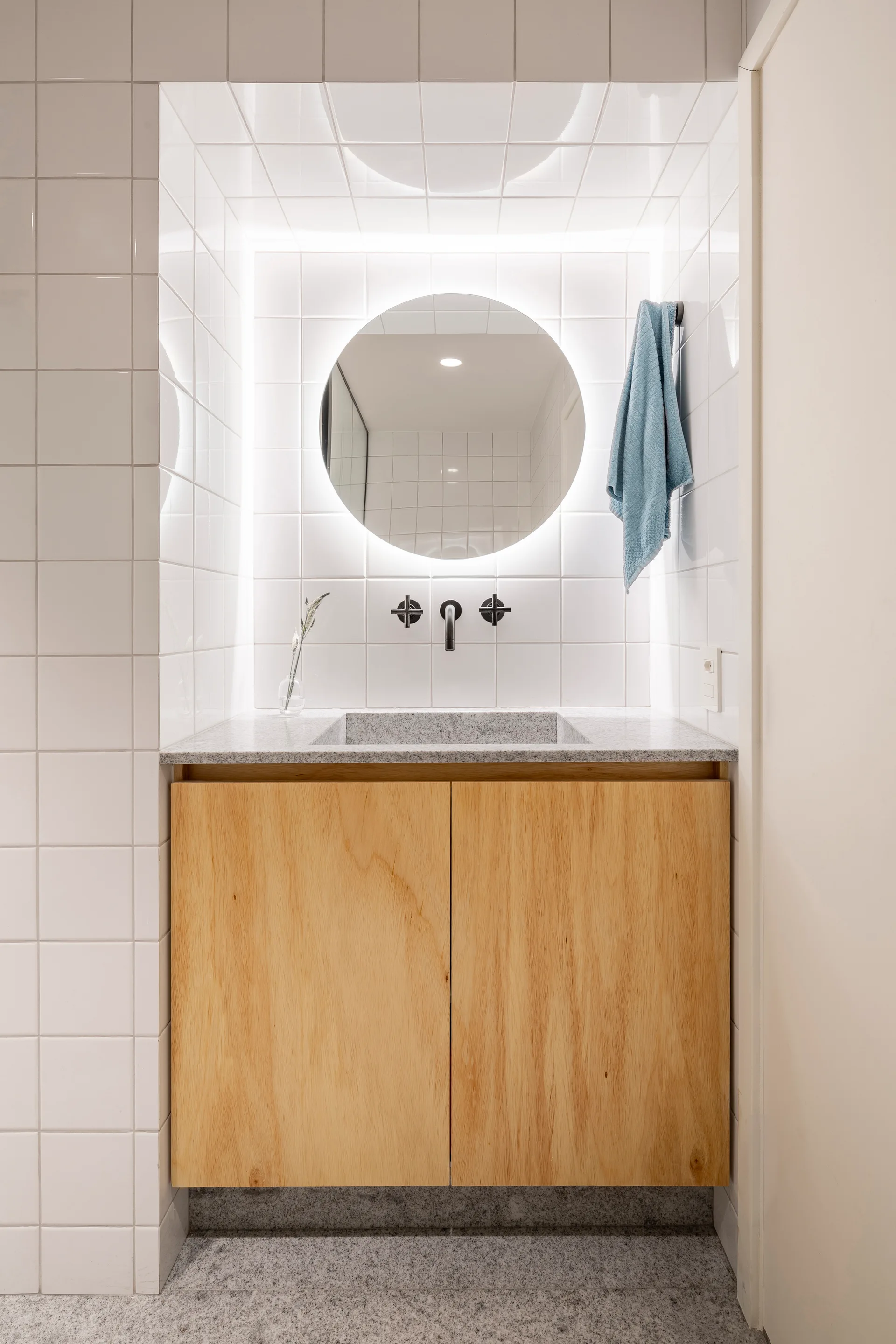 A recessed wood vanity features a stone countertop, a black faucet, and a round backlit mirror on a white square-tiled wall.