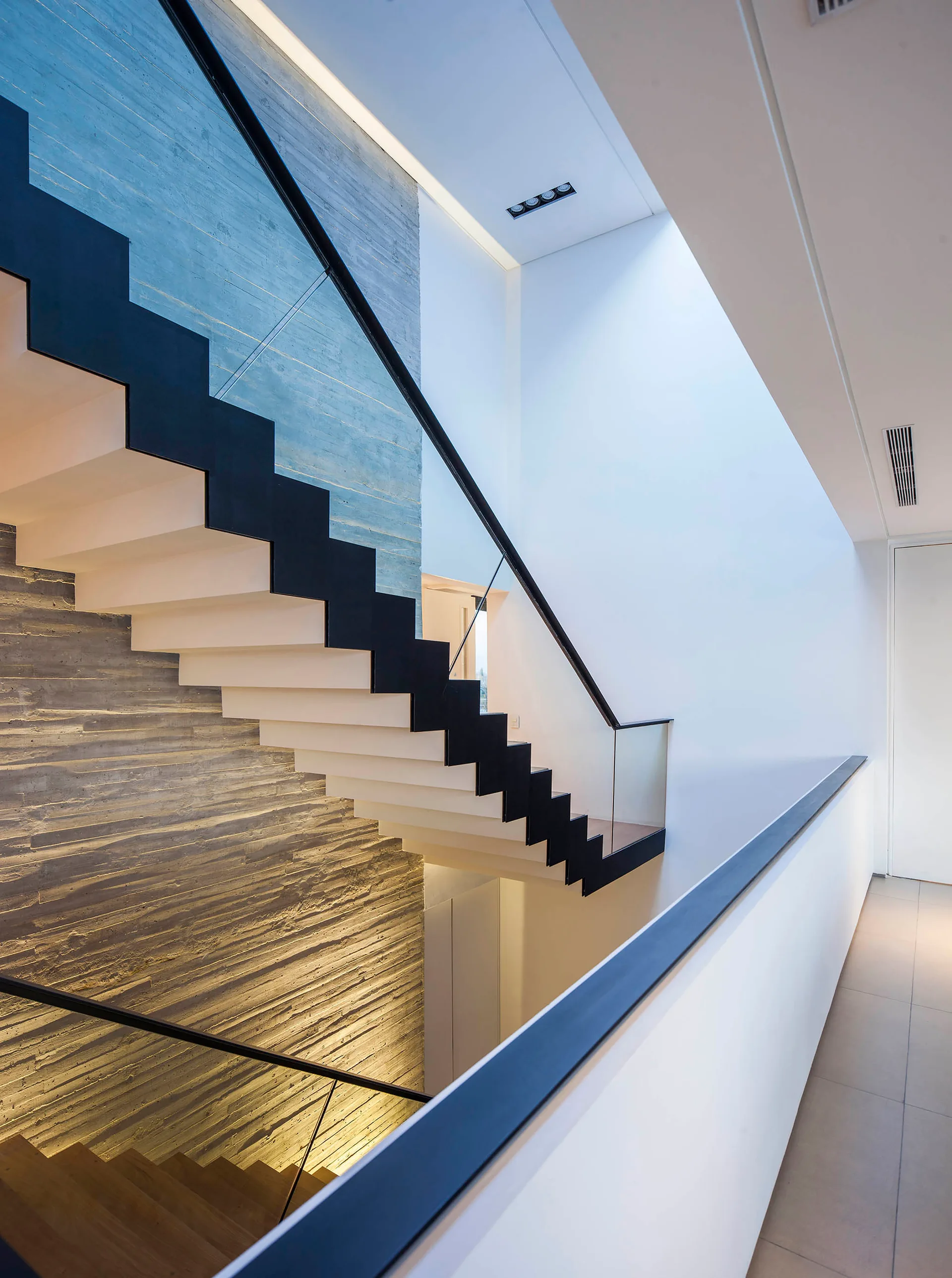 A modern open-riser staircase with a black frame and glass railing stands against a tall, textured board-formed concrete wall.