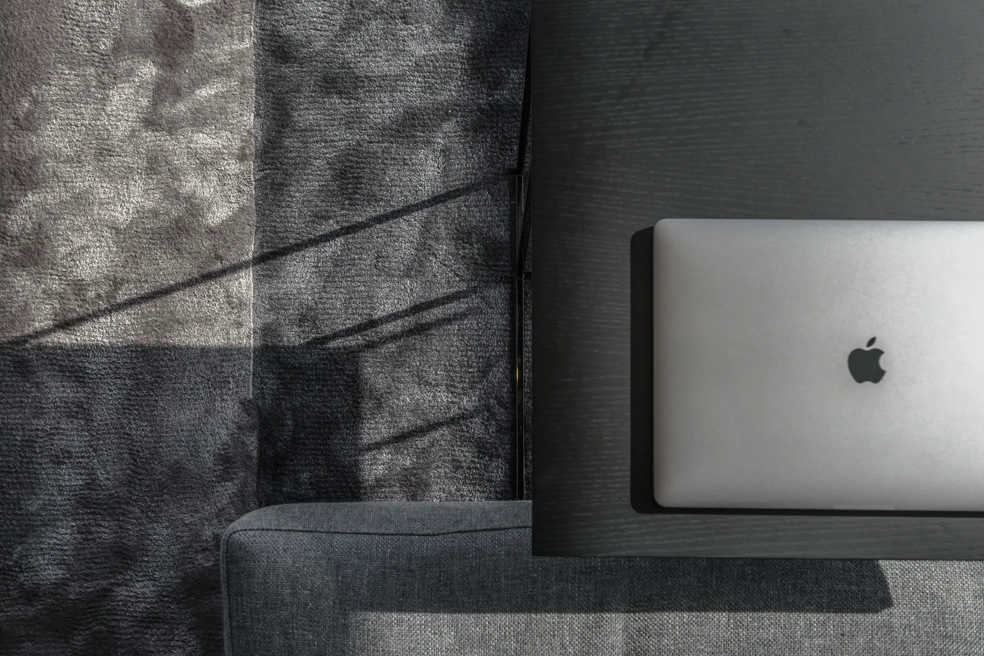 A top-down view of a silver laptop on a dark wood desk next to a grey upholstered sofa and a textured rug with striped shadows.