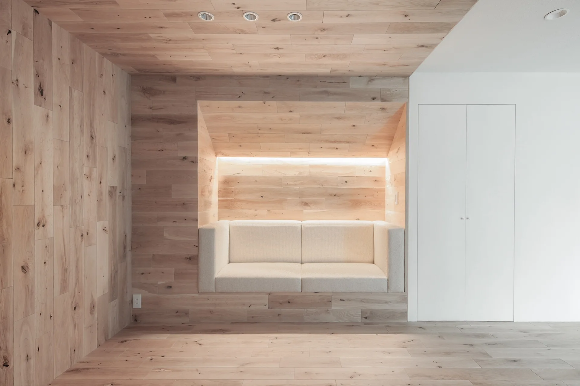 A minimalist room featuring a beige sofa recessed into a light wood-paneled wall alcove beside a plain white closet door.