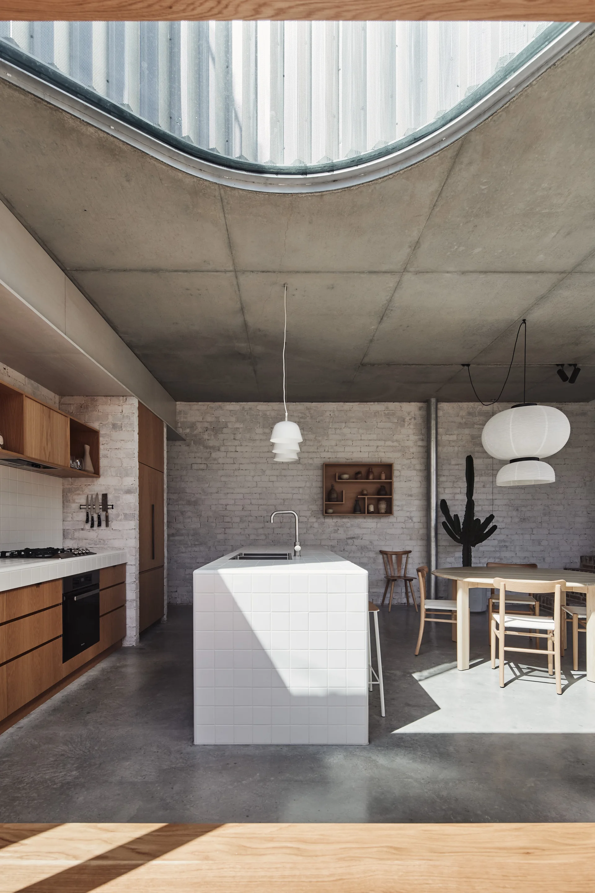A minimalist kitchen features a white tiled island, wooden cabinetry, and a circular skylight set in a raw concrete ceiling.