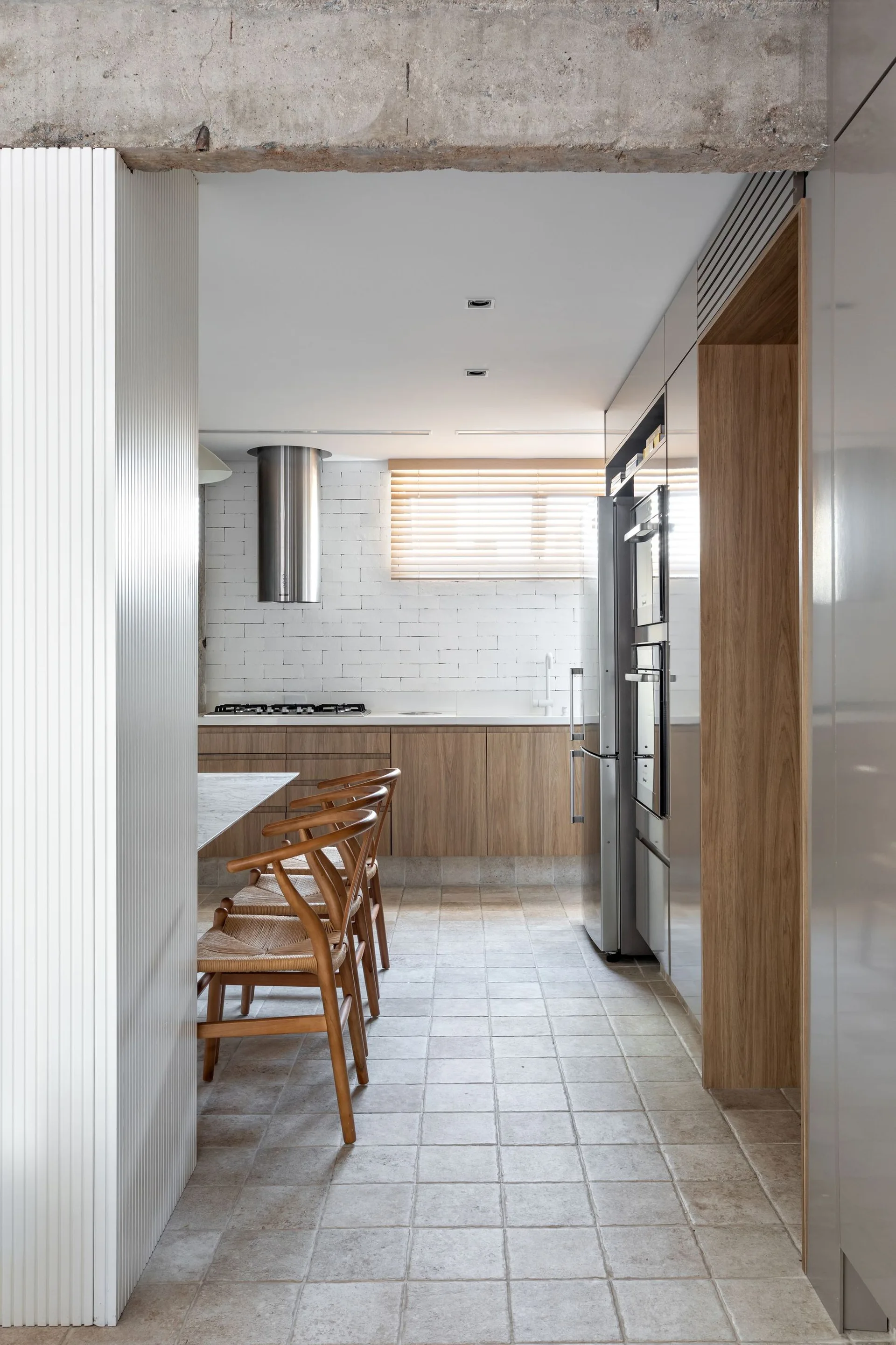 A modern kitchen featuring wooden cabinets, a white brick wall, an exposed concrete beam overhead, and woven wood chairs.