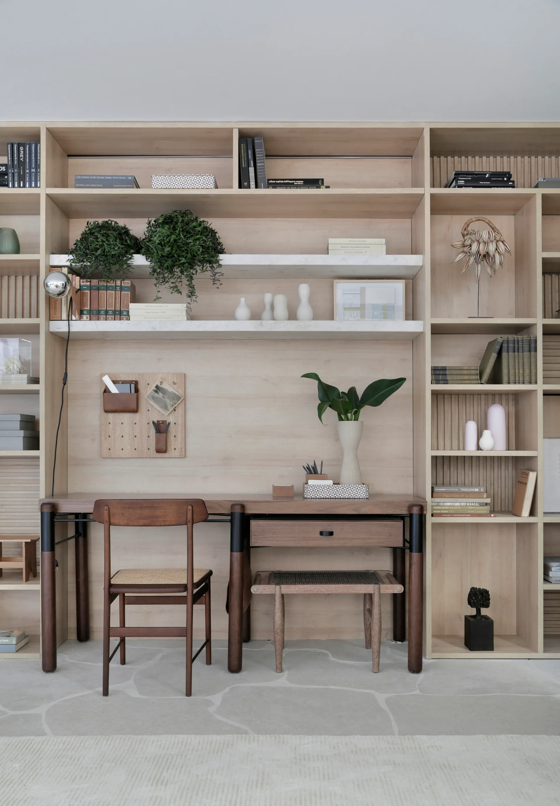 A built-in light wood shelving unit with an integrated desk, marble shelves, a wooden desk chair, and light stone tile flooring.