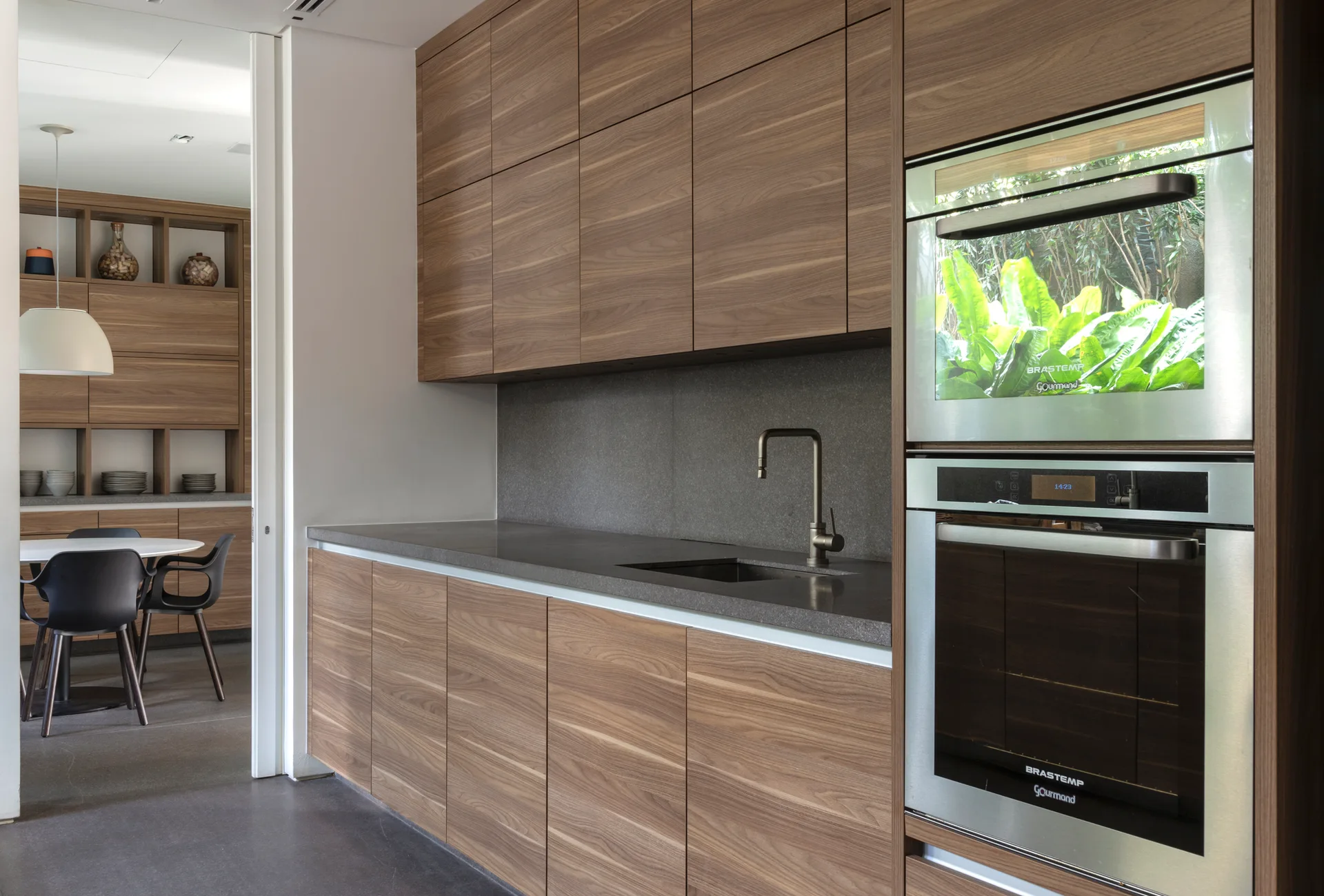 A modern kitchen featuring flat-panel wood cabinetry, integrated stainless steel ovens, and a dark grey stone countertop and backsplash.
