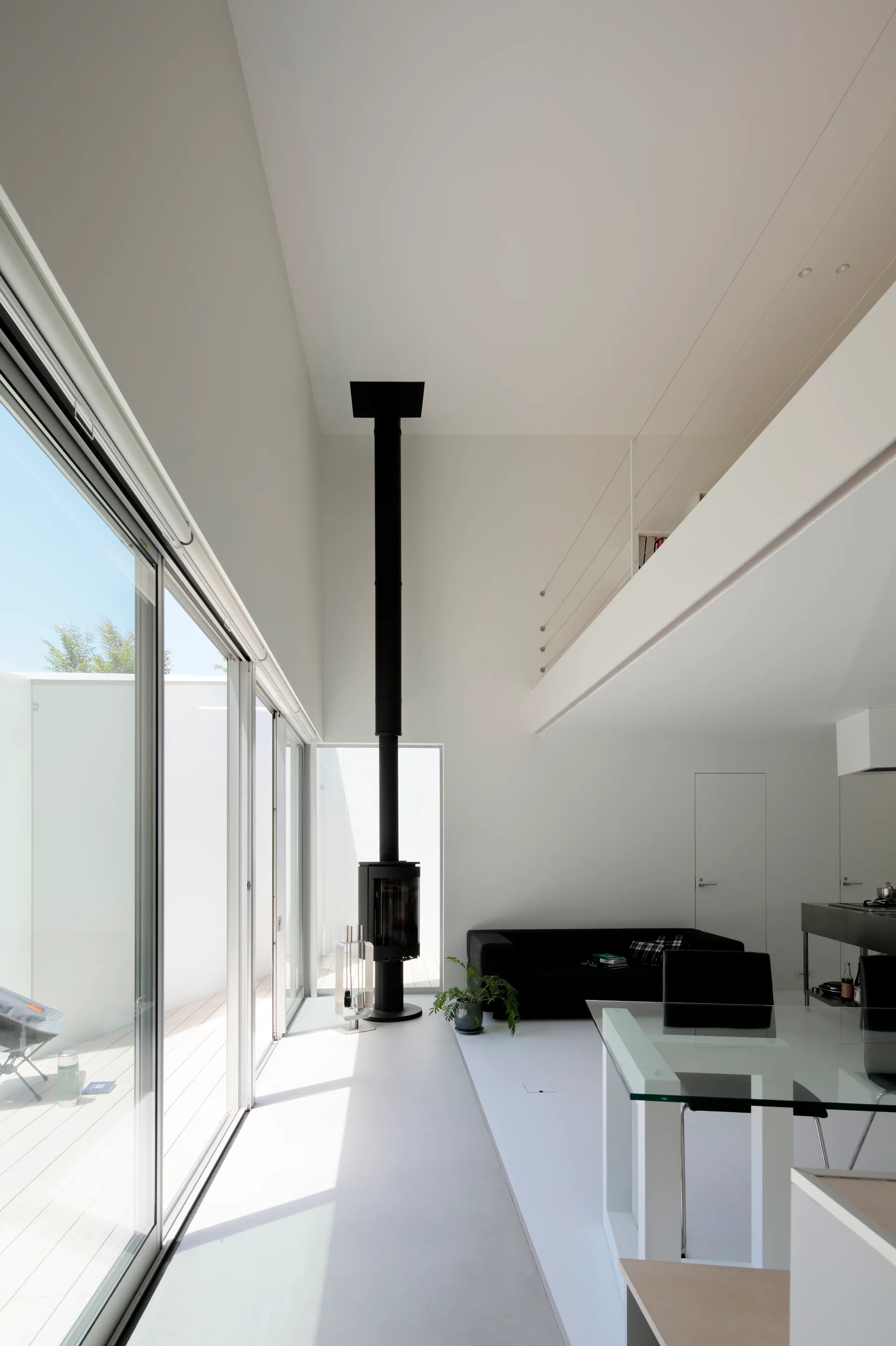 A minimalist white living room features a tall black fireplace, a glass-topped dining table, and large sliding glass doors beneath a mezzanine.