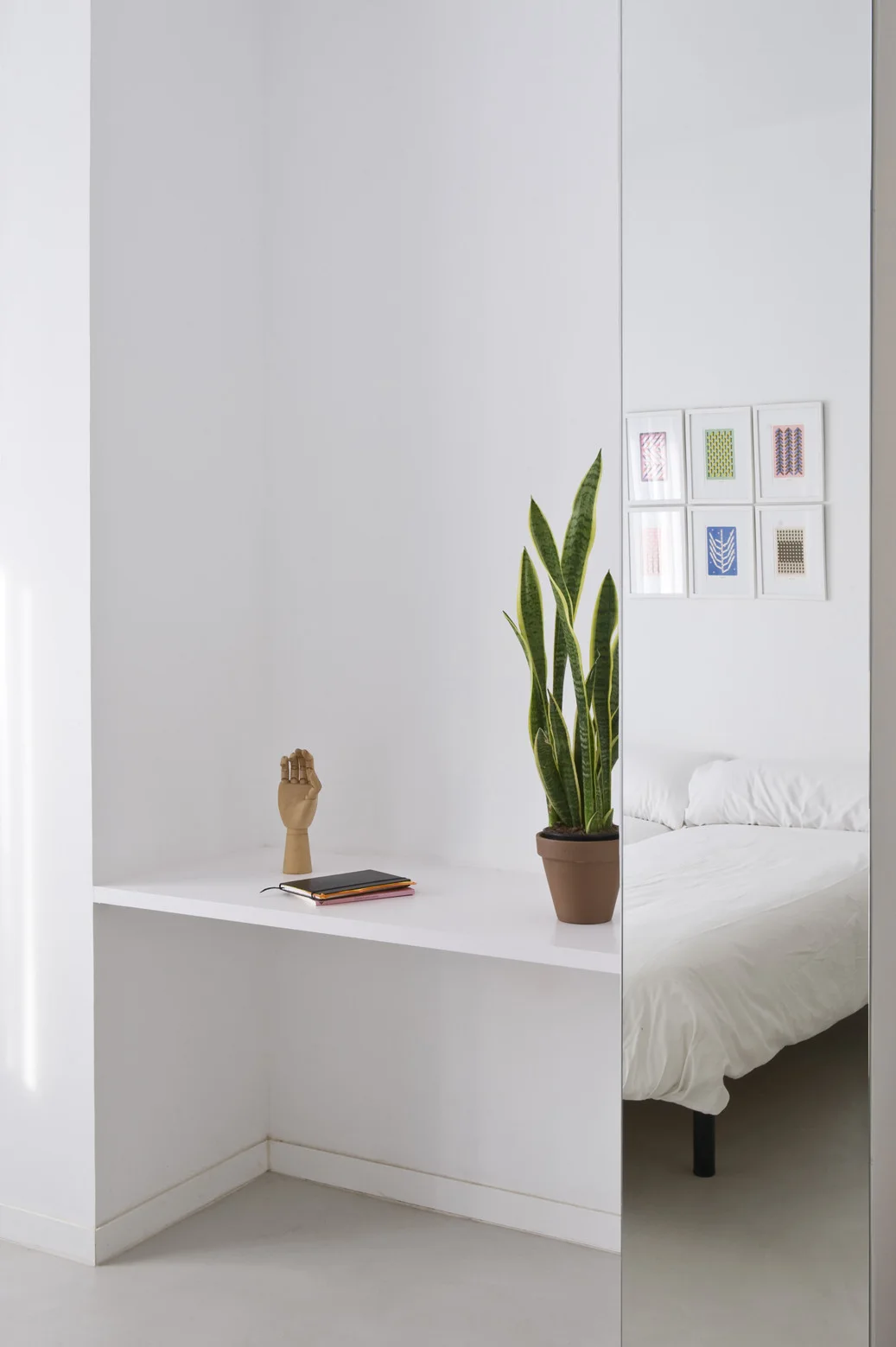 A minimalist white bedroom featuring a built-in floating desk shelf with a potted plant, alongside a tall mirror reflecting a bed and framed artwork.