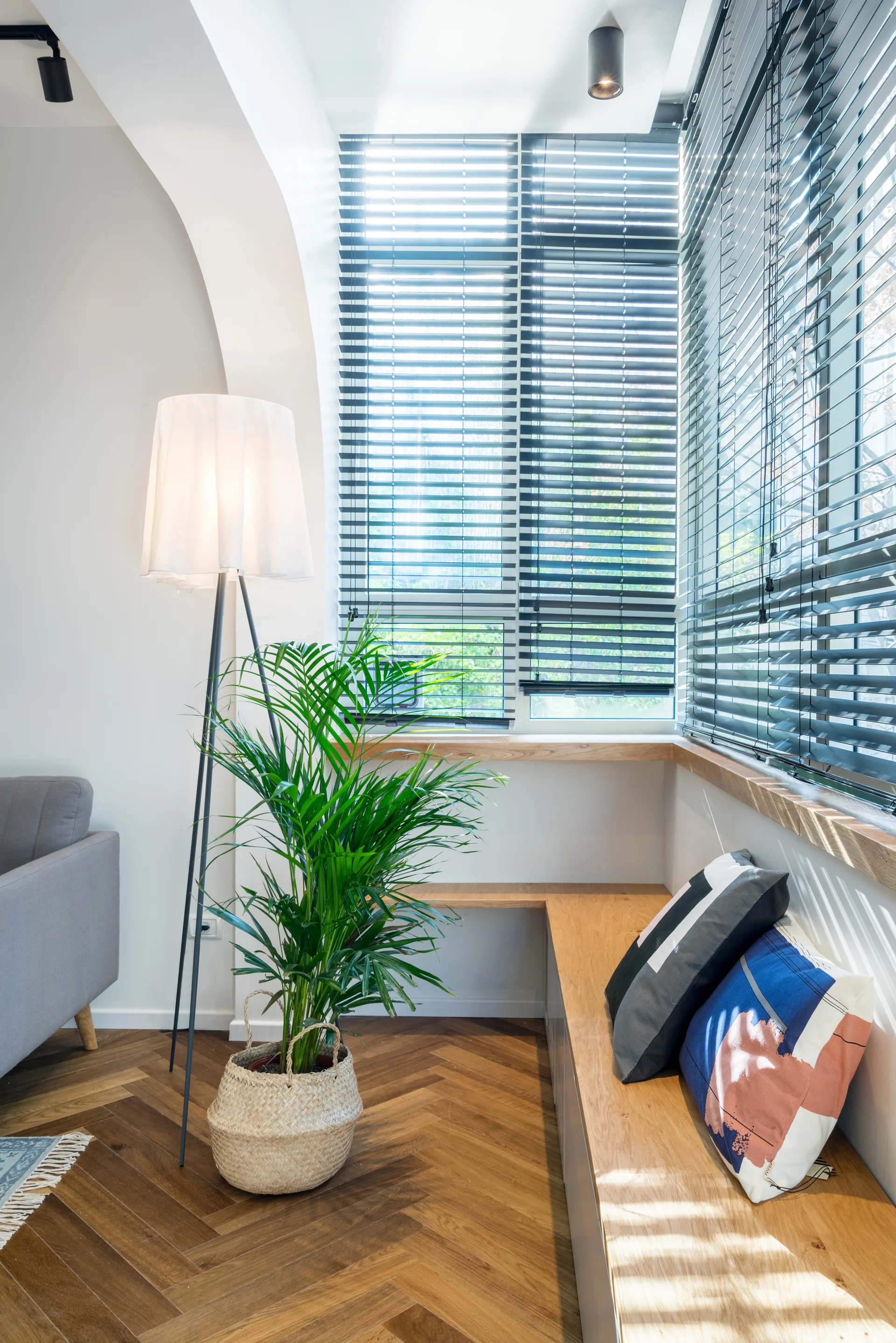 A built-in wooden corner bench with throw pillows sits beneath large windows with dark blinds in a room featuring herringbone wood floors.