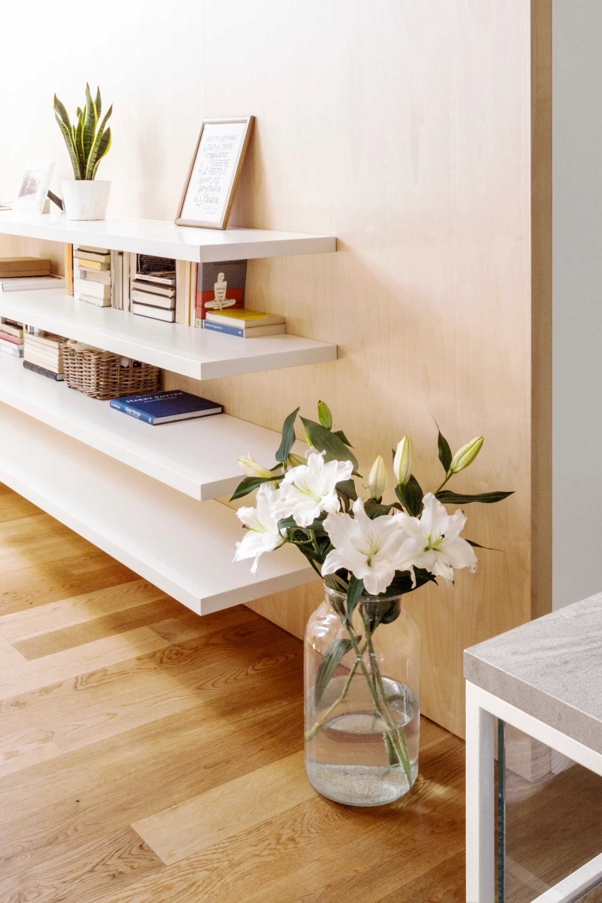White floating shelves styled with books and plants on a light wood wall, with a glass vase of lilies on the wooden floor.