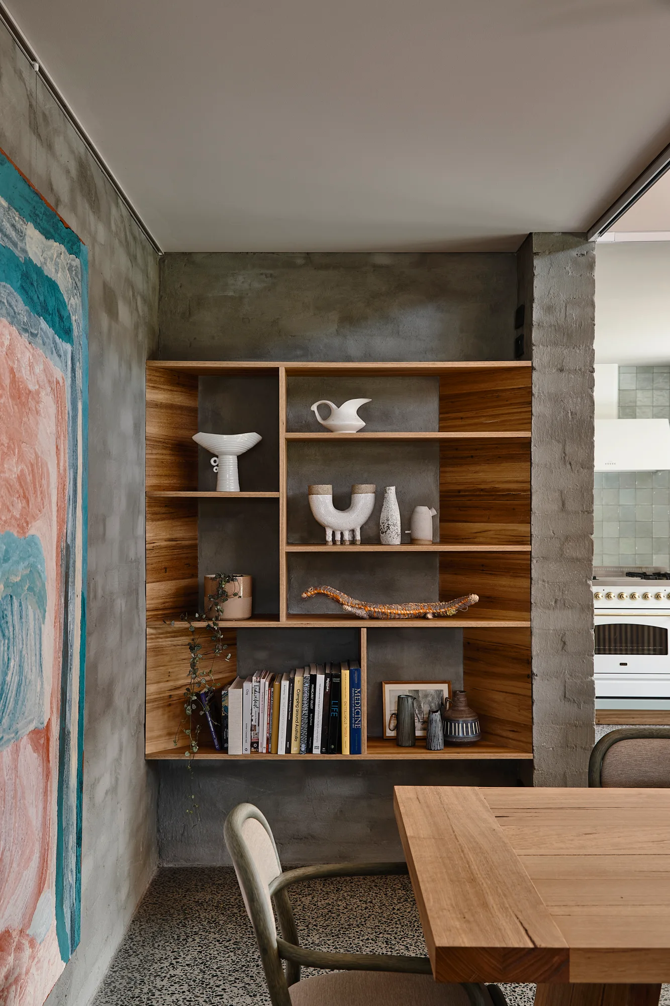 A recessed oak shelving unit is built into a textured concrete wall beside a wooden dining table and chair in a room with speckled terrazzo floors.