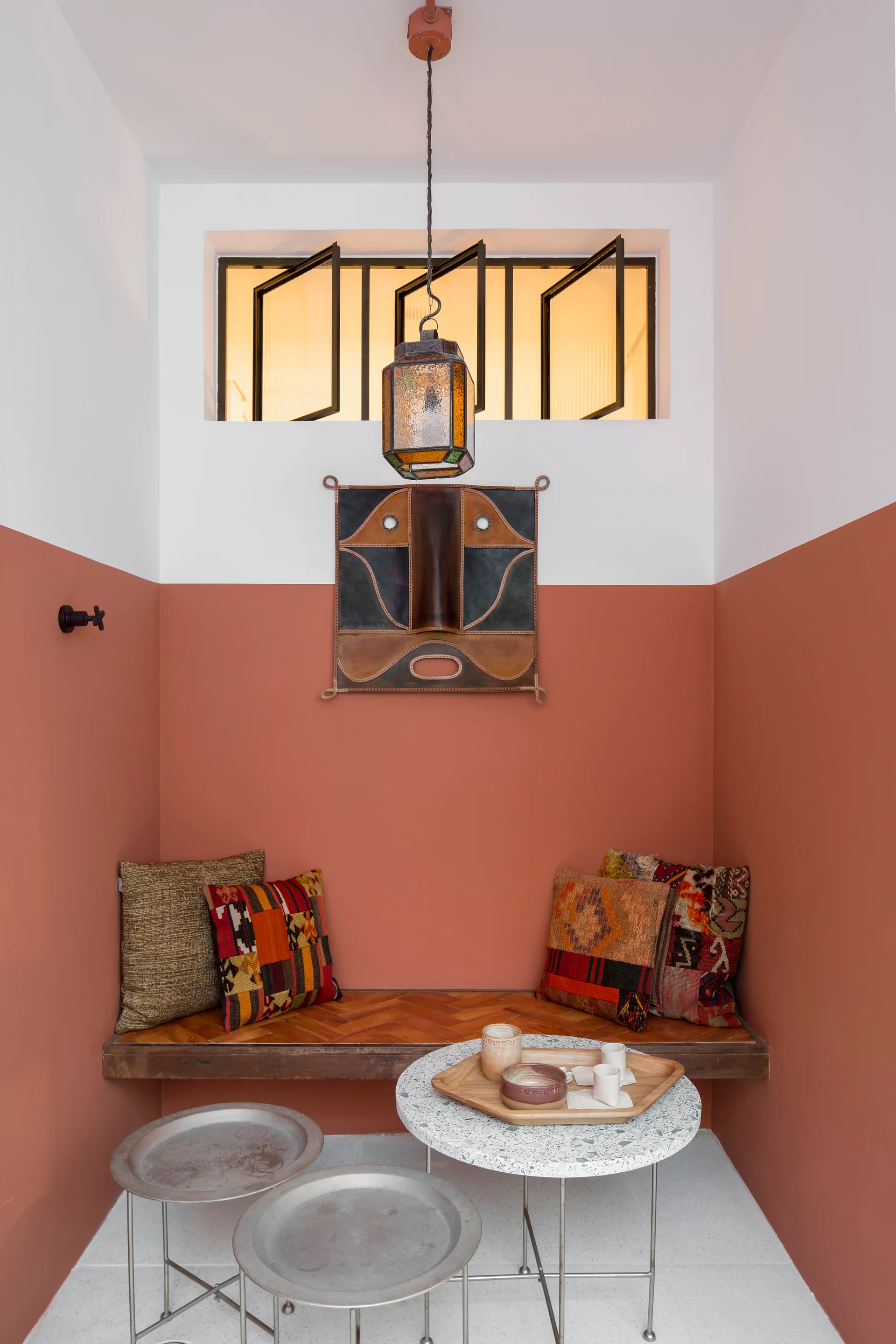 A cozy seating nook features a two-toned white and terracotta wall behind a built-in wooden bench, accented by patterned cushions and a terrazzo table.