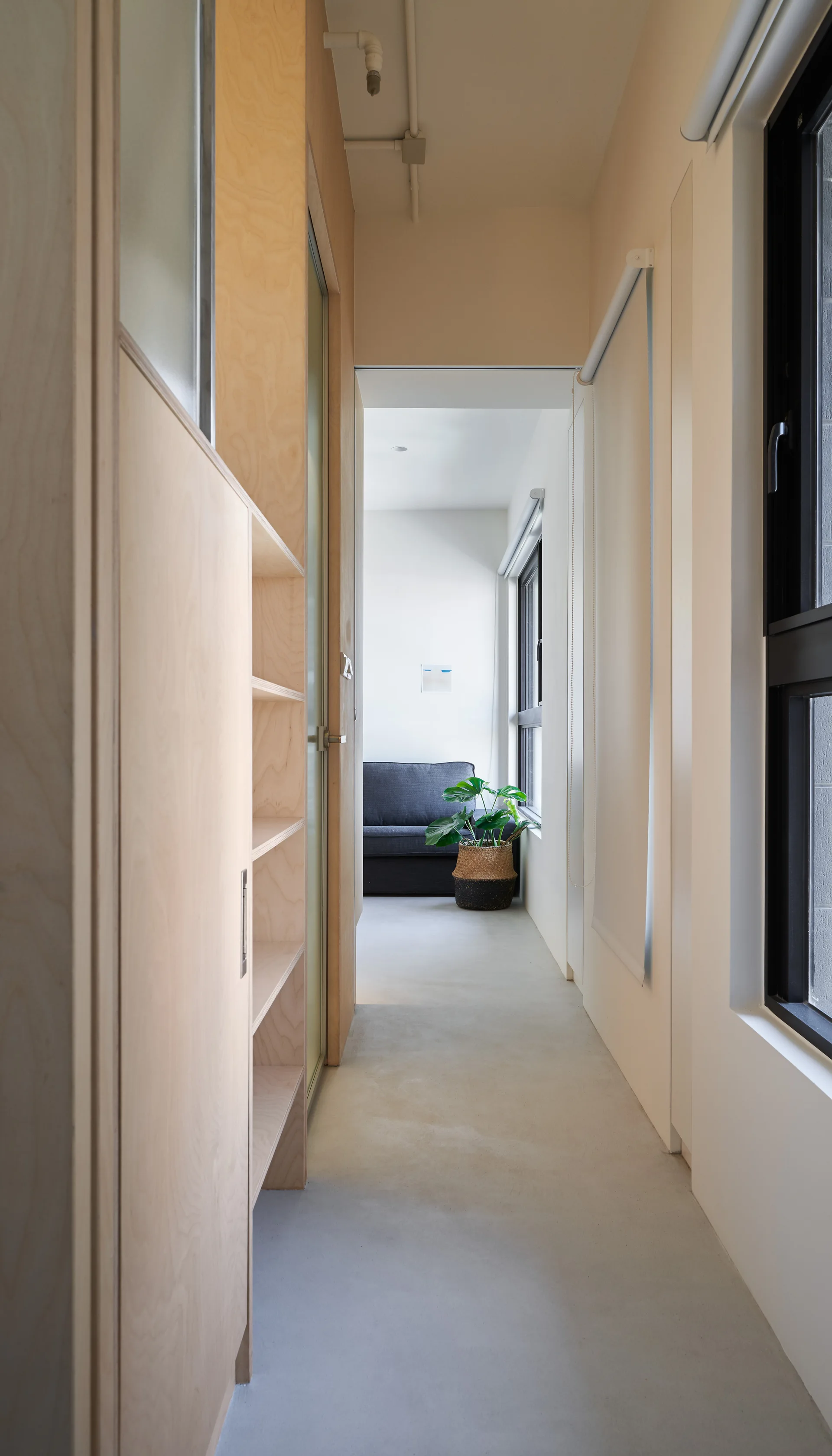 A narrow residential hallway featuring built-in light plywood cabinetry on the left and black-framed windows on the right, leading to a living room.
