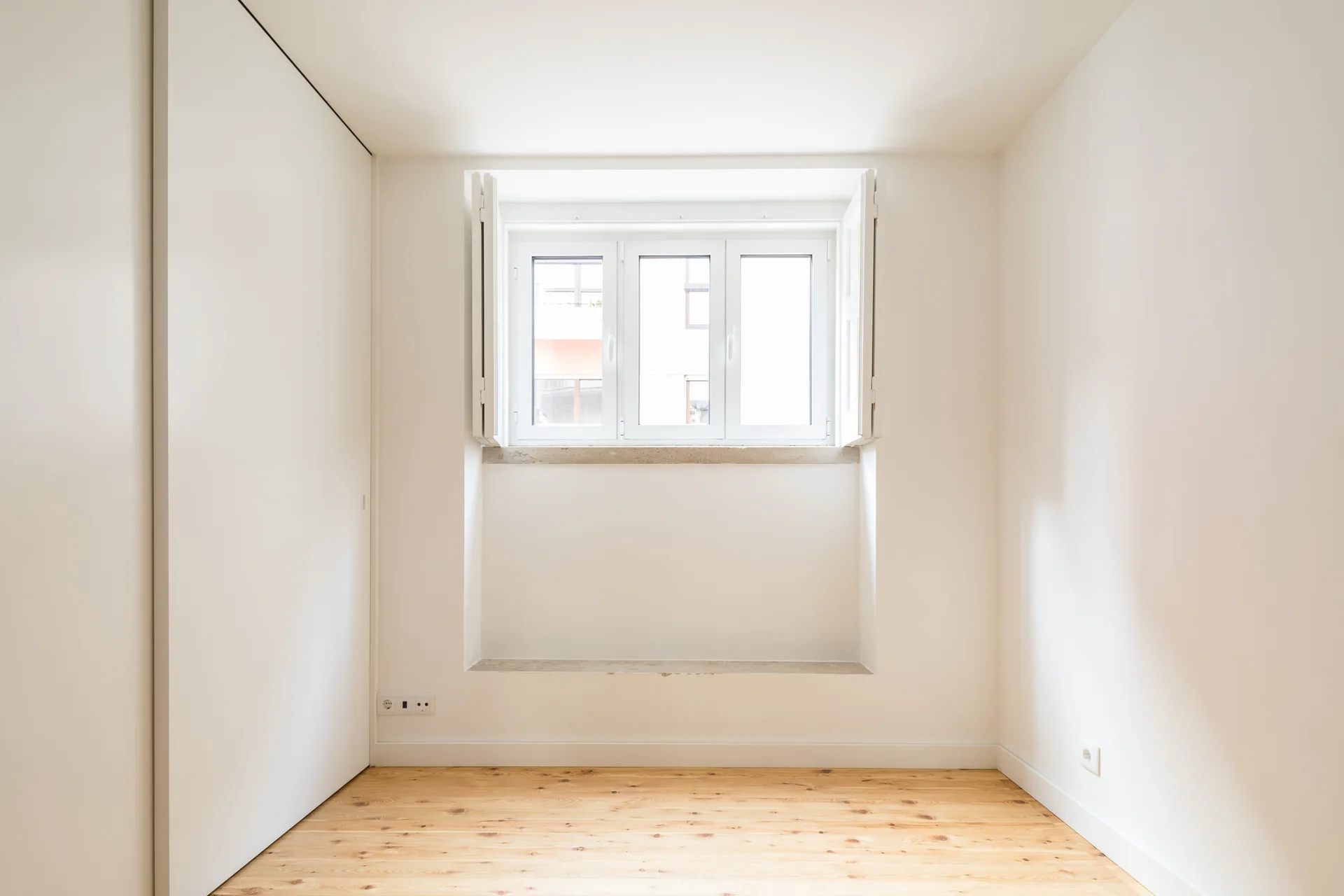 An empty minimalist room featuring natural wood floors, warm white walls, and a central window with open interior shutters.