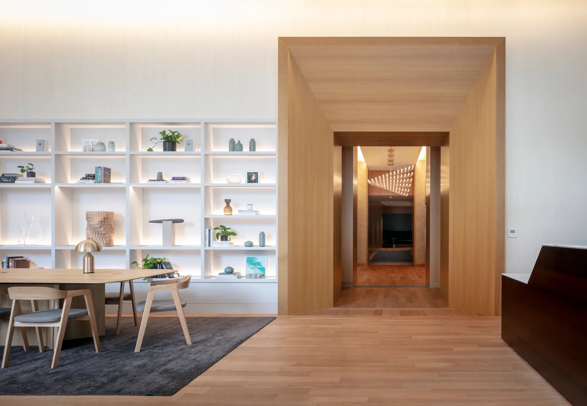 An office lobby features a large wooden archway, a minimalist table with chairs, and white built-in shelves illuminated by integrated lighting.